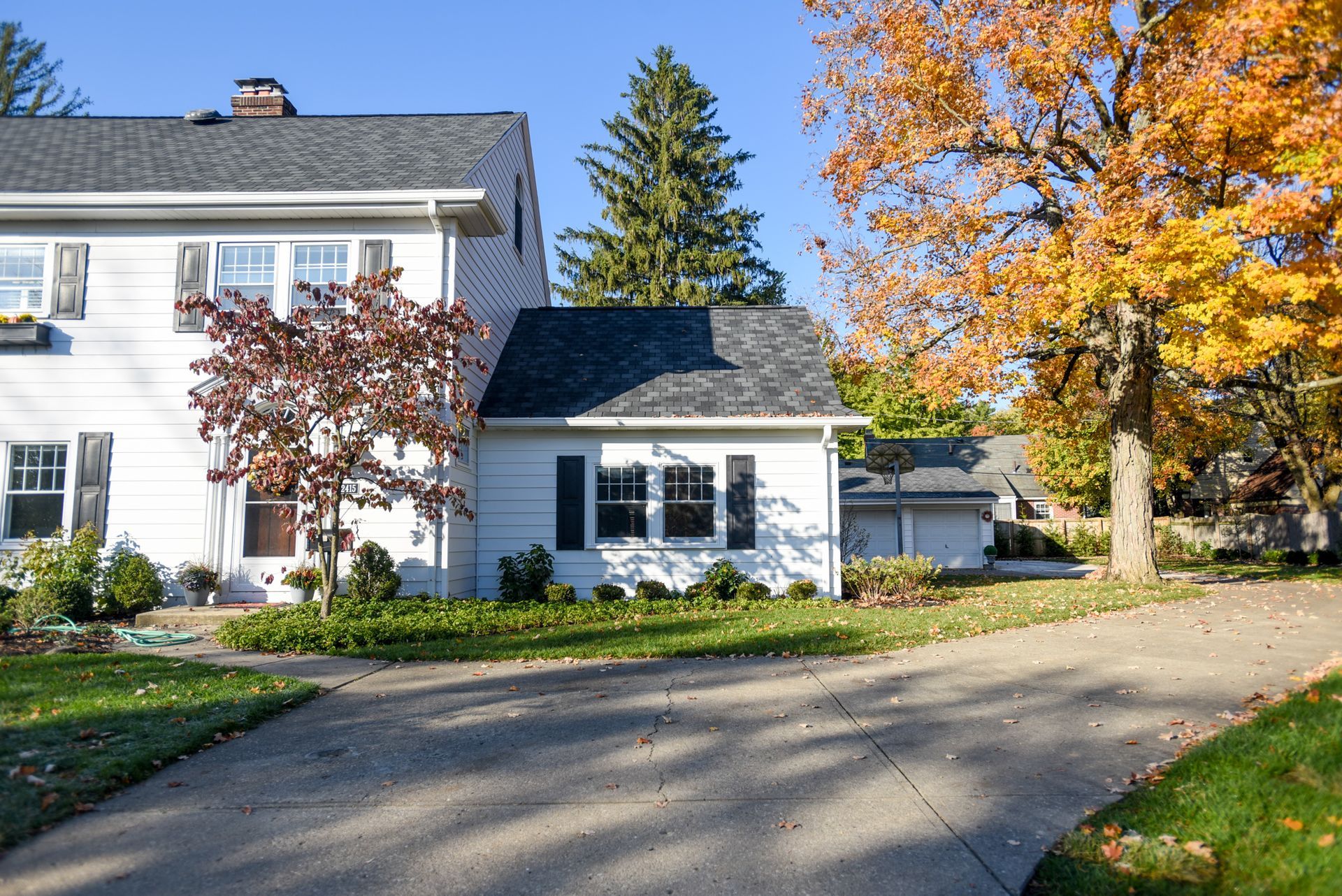 White house with black shutters and autumn trees beside a curved driveway.