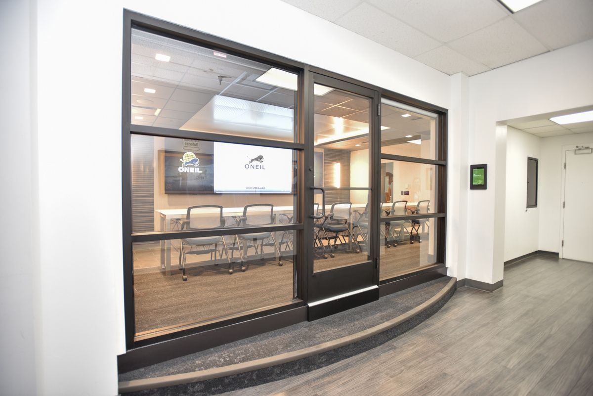 Glass conference room with long table, office chairs, wall screens, and curved hallway outside.