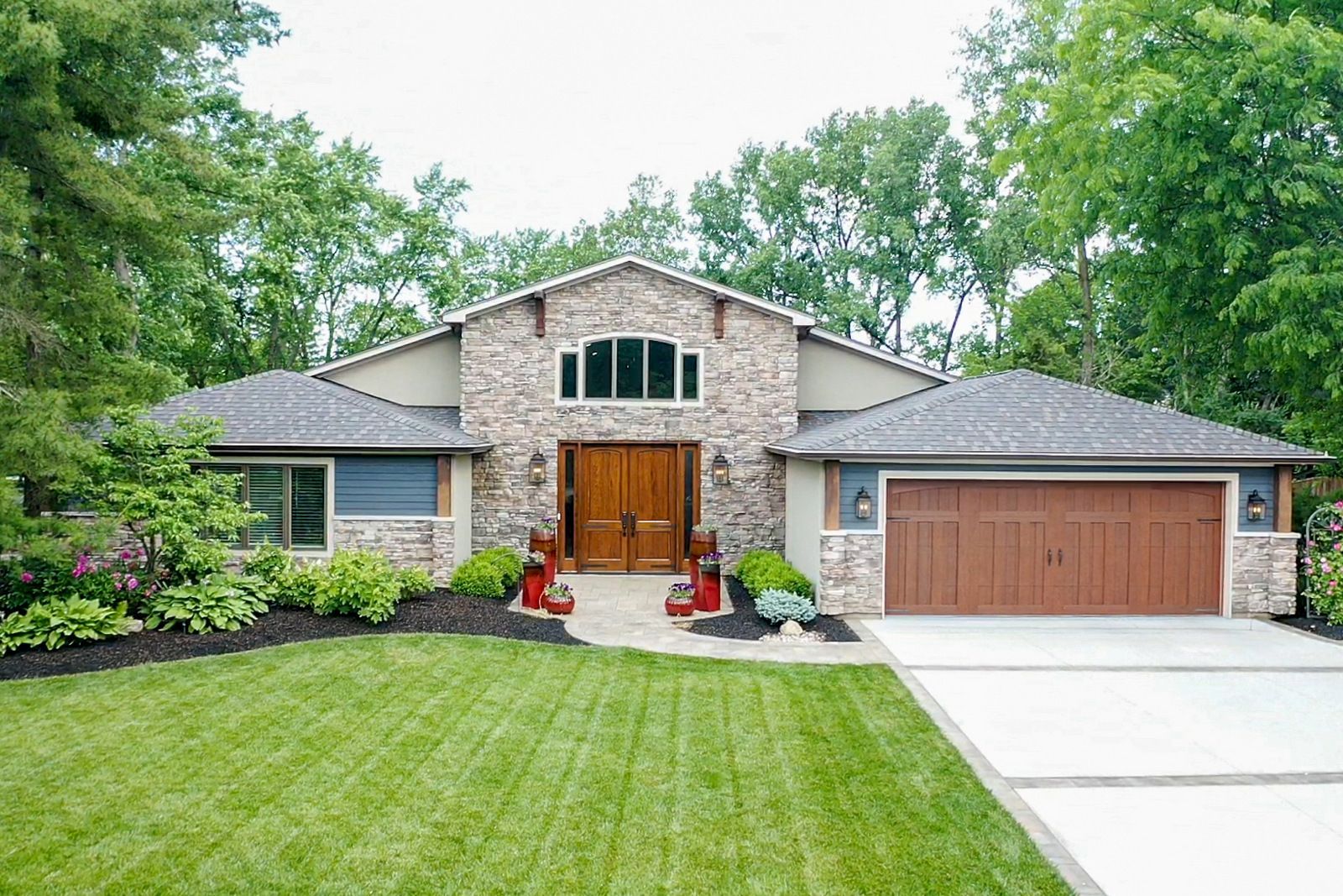 Suburban stone house with a wooden garage door, green lawn, and tree-lined front yard