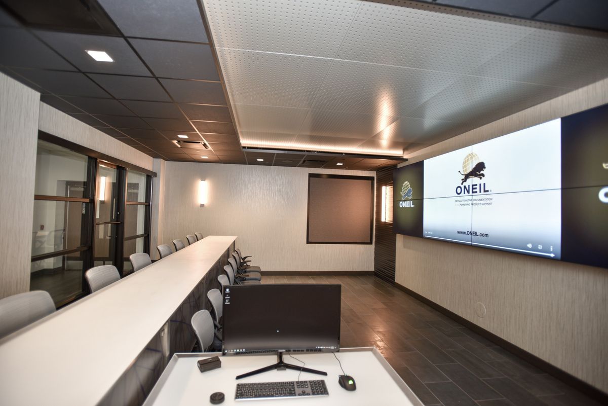 Empty conference room with a long white table, wall-mounted screen, and dark carpet flooring.