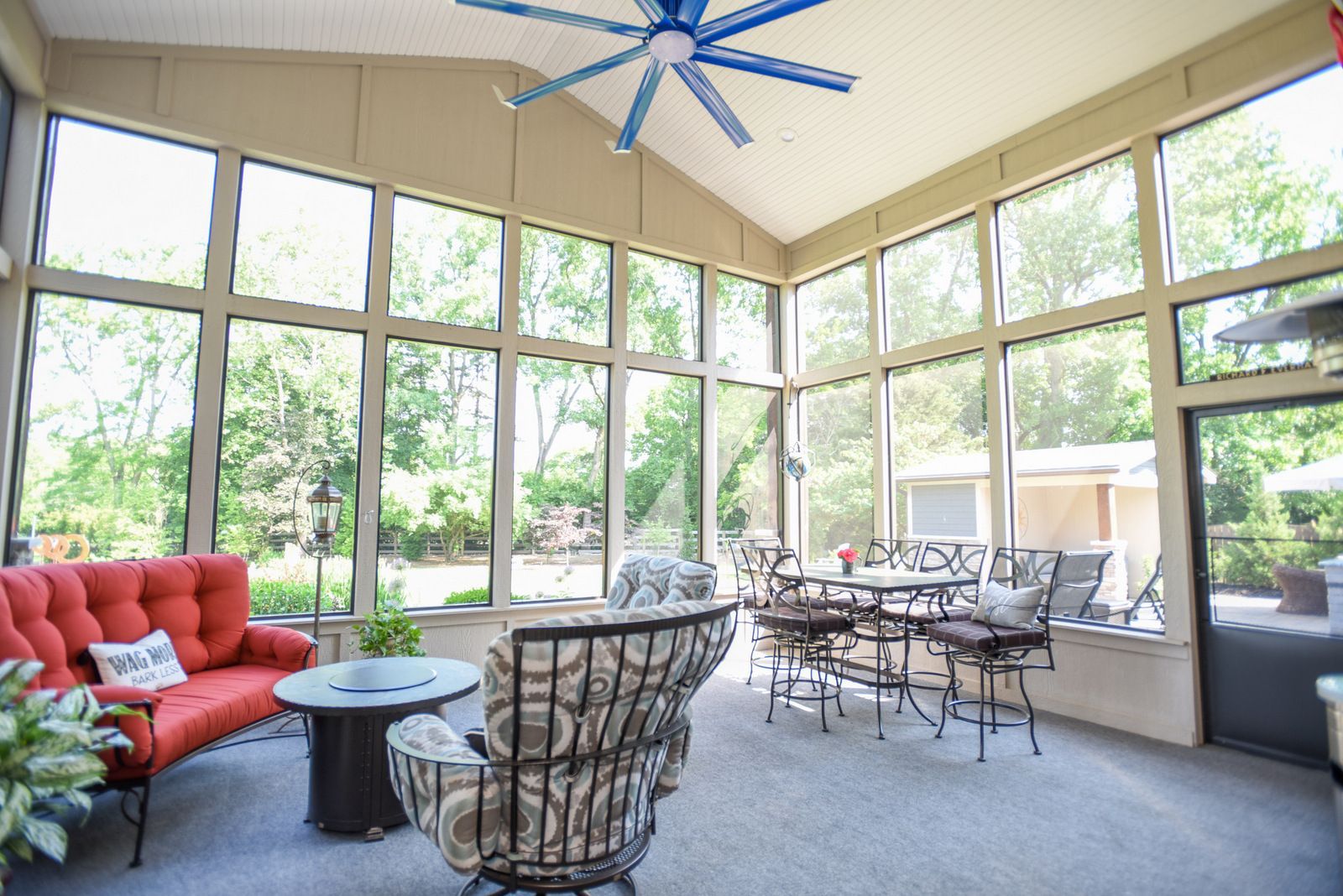 Bright sunroom with large windows, red sofa, wicker chairs, and a blue ceiling fan.