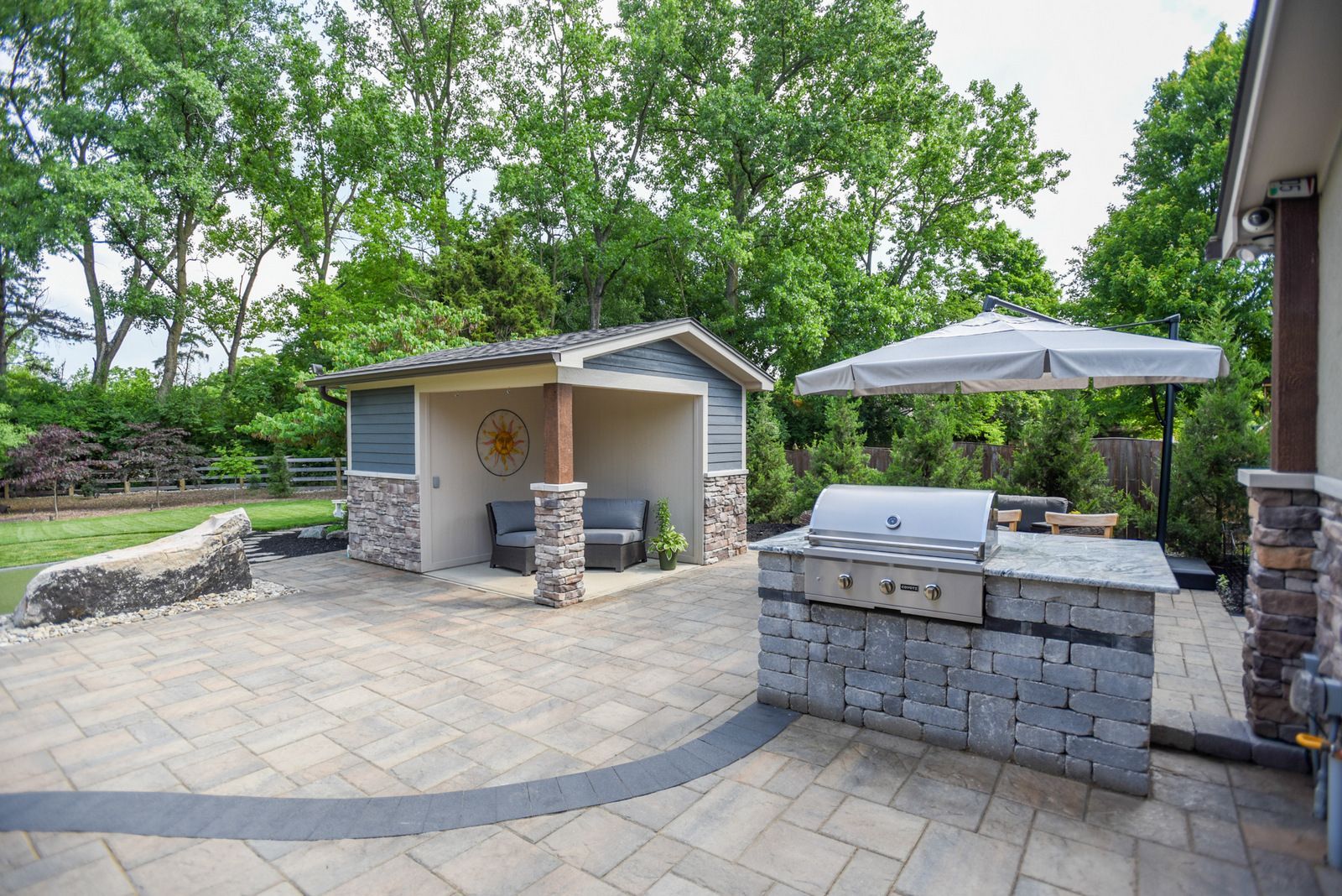 Outdoor stone patio with built-in grill, small shelter, umbrella, and trees surrounding the area.