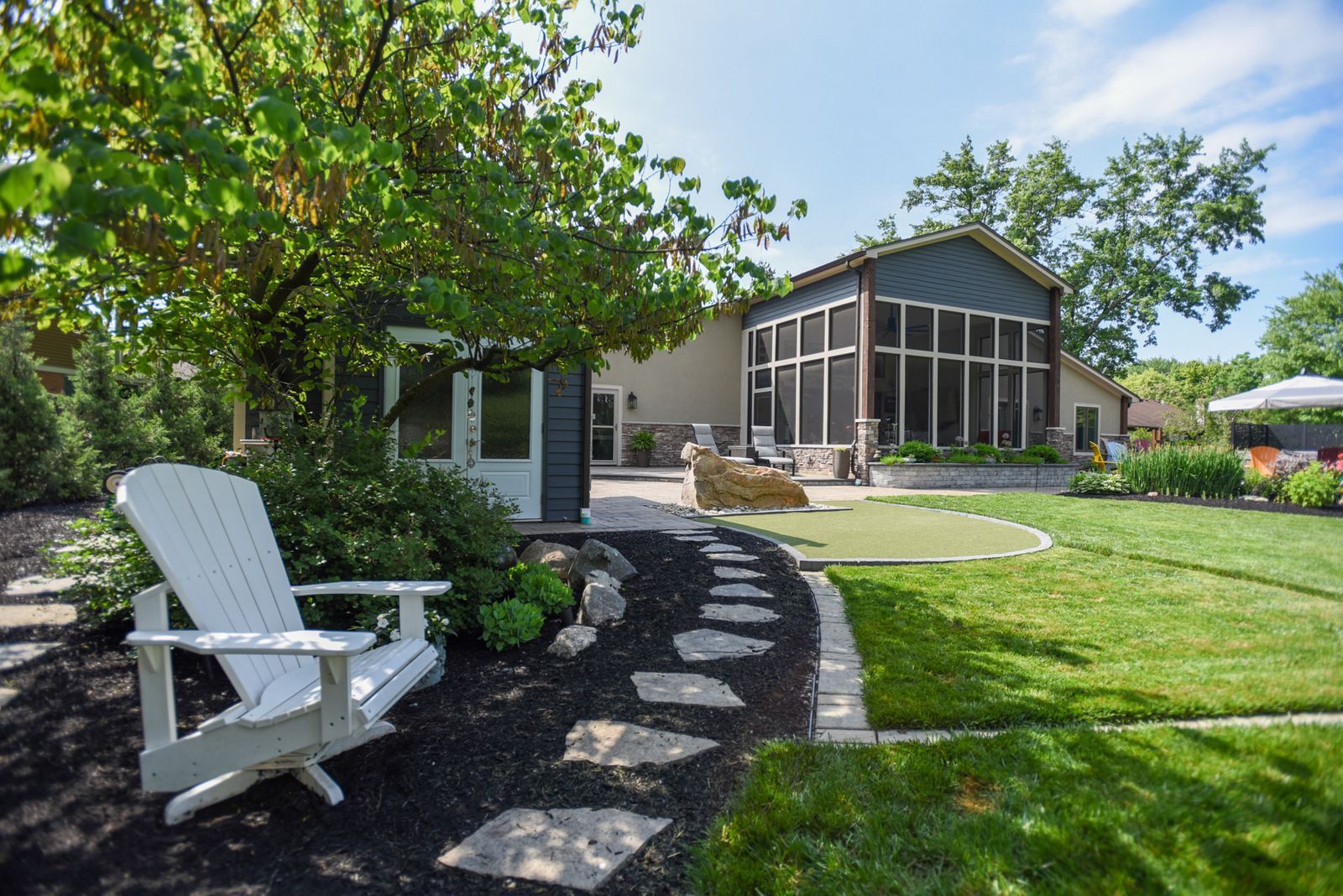 Backyard patio with white Adirondack chair, curved stone path, lush lawn, and screened porch on a sunny day.