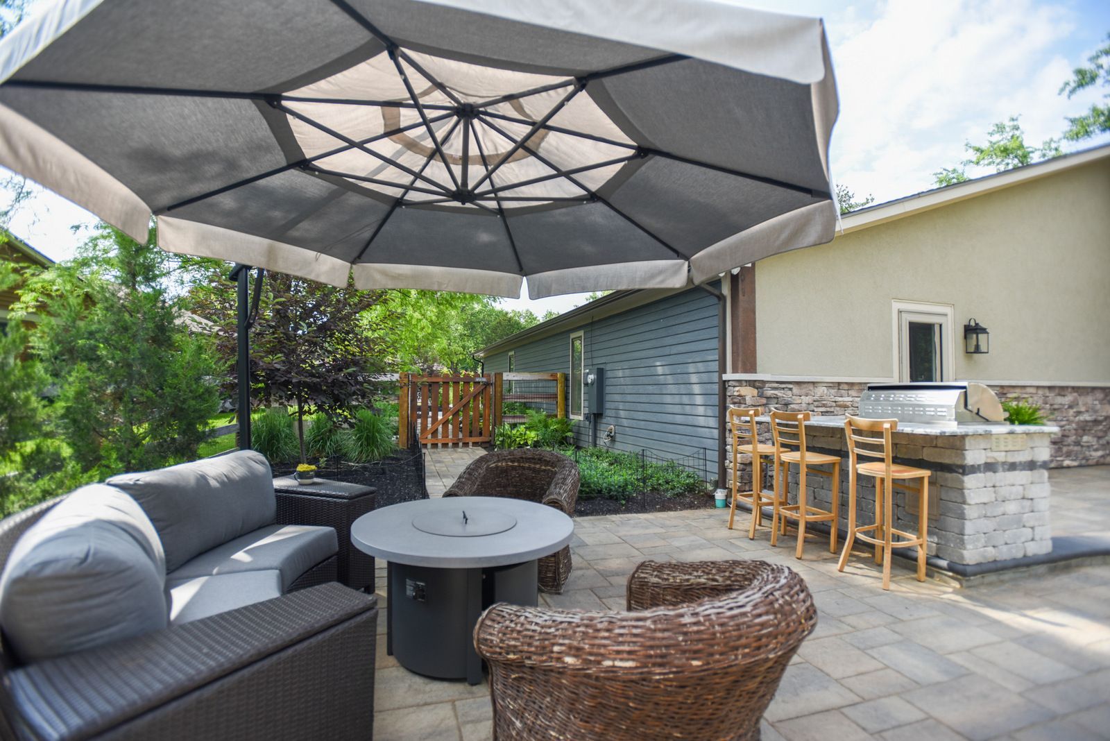 Patio seating area with gray sofa, wicker chairs, large umbrella, and outdoor bar beside a house.
