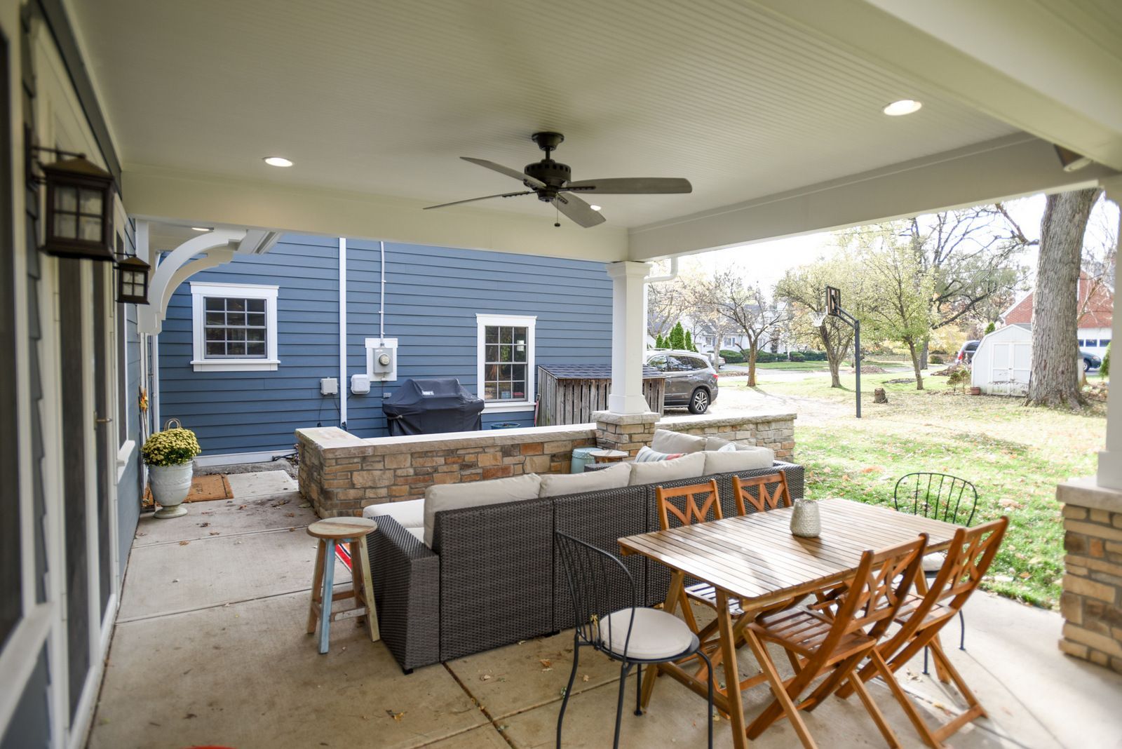 Covered patio with wicker furniture, dining table, and stone fire pit beside a blue house and grassy yard.