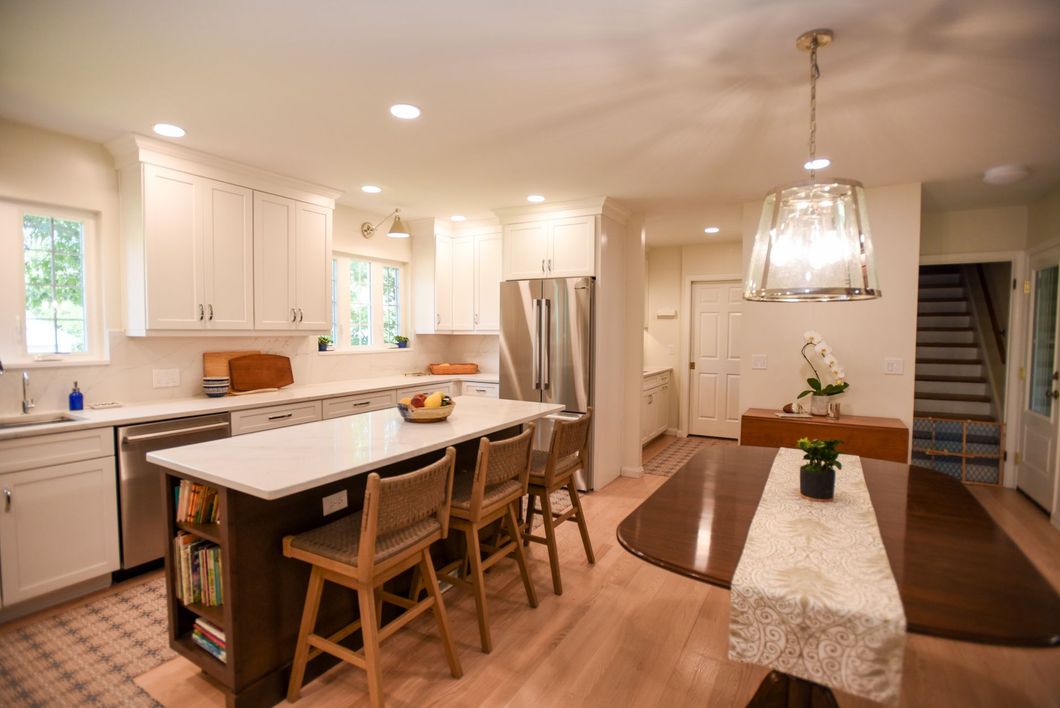 Bright modern kitchen and dining area with island, wood floors, and staircase in the background