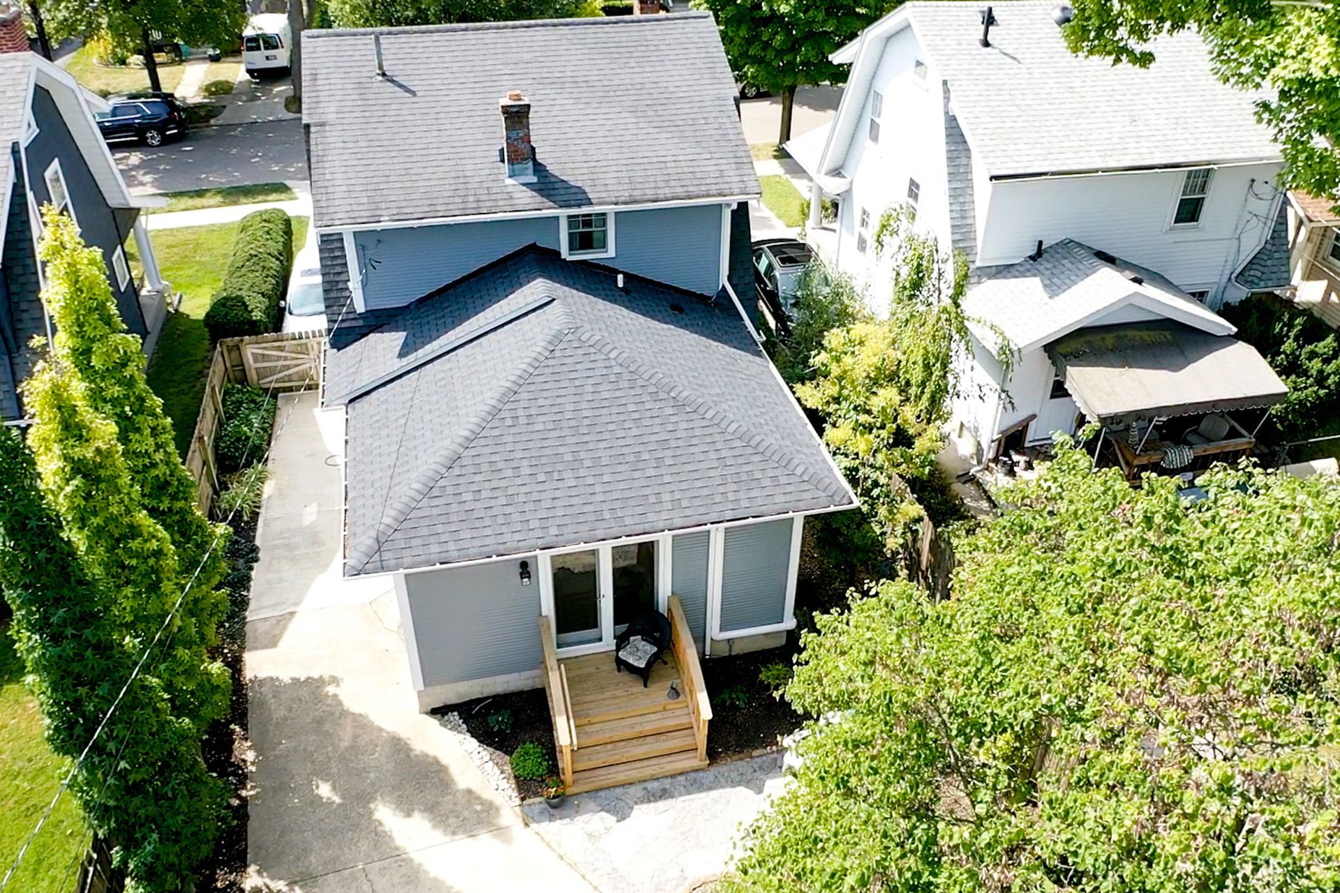 Aerial view of a small gray house with a front porch in a leafy suburban neighborhood.