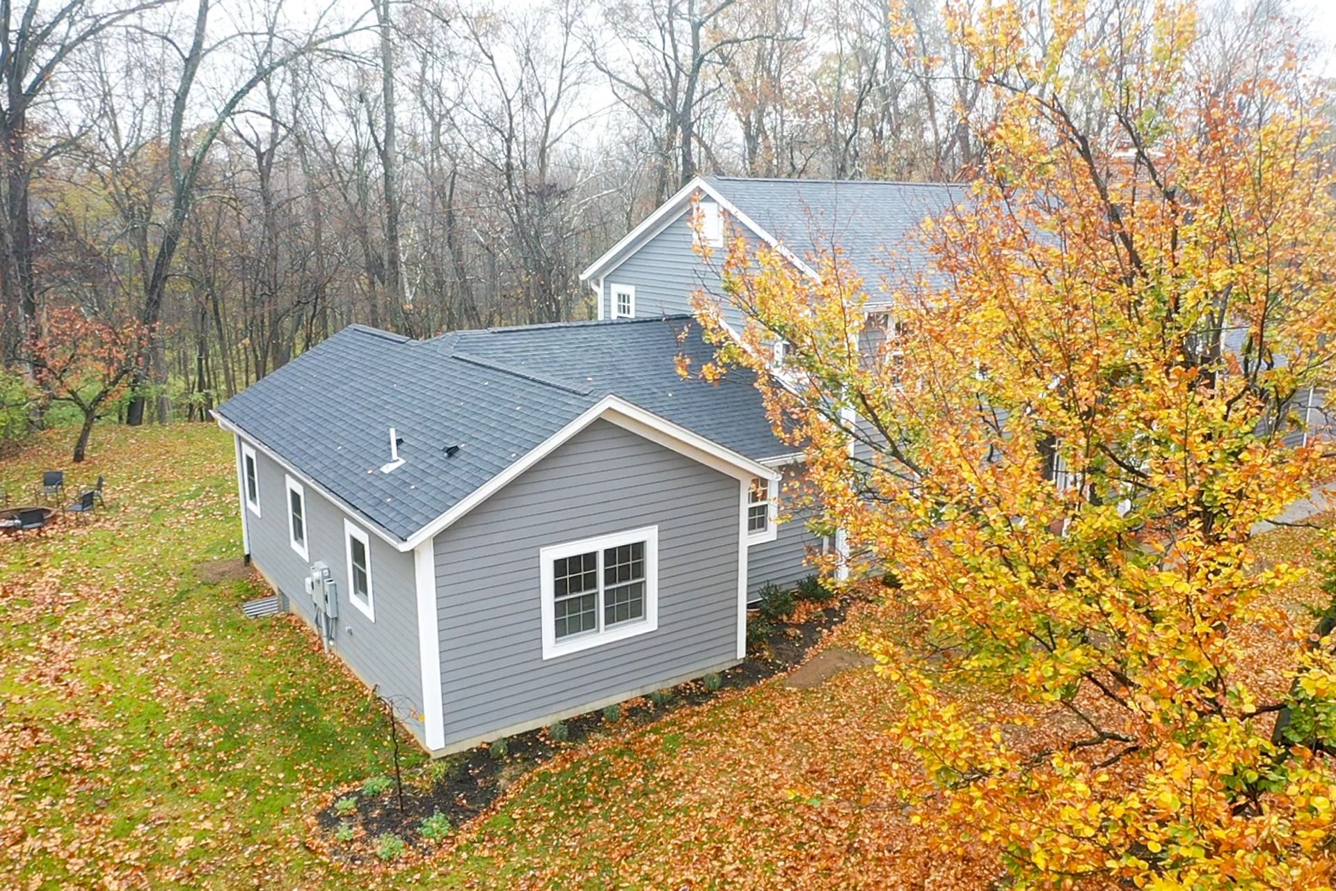 Gray house with blue roof beside an autumn tree and fallen leaves in a wooded yard.