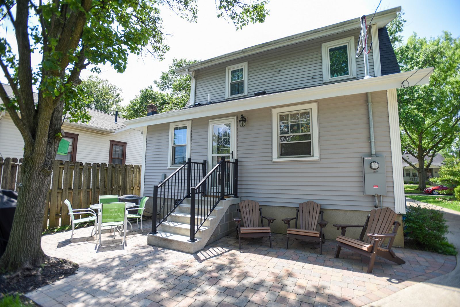 Two-story beige house with front porch, steps, and patio chairs under trees.