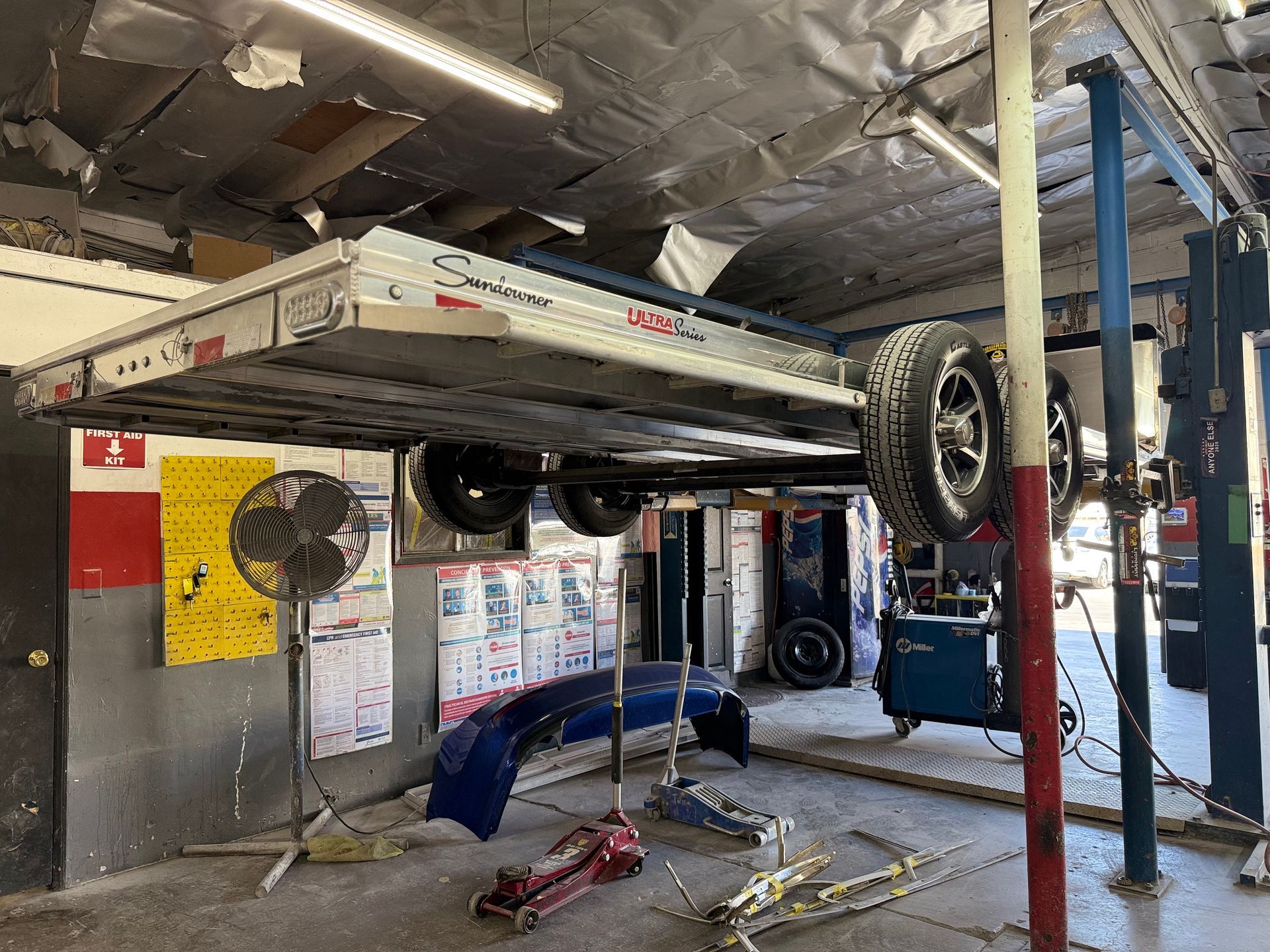 Car on a lift in an auto repair shop. Tires, bumper, tools, and a fan are visible.