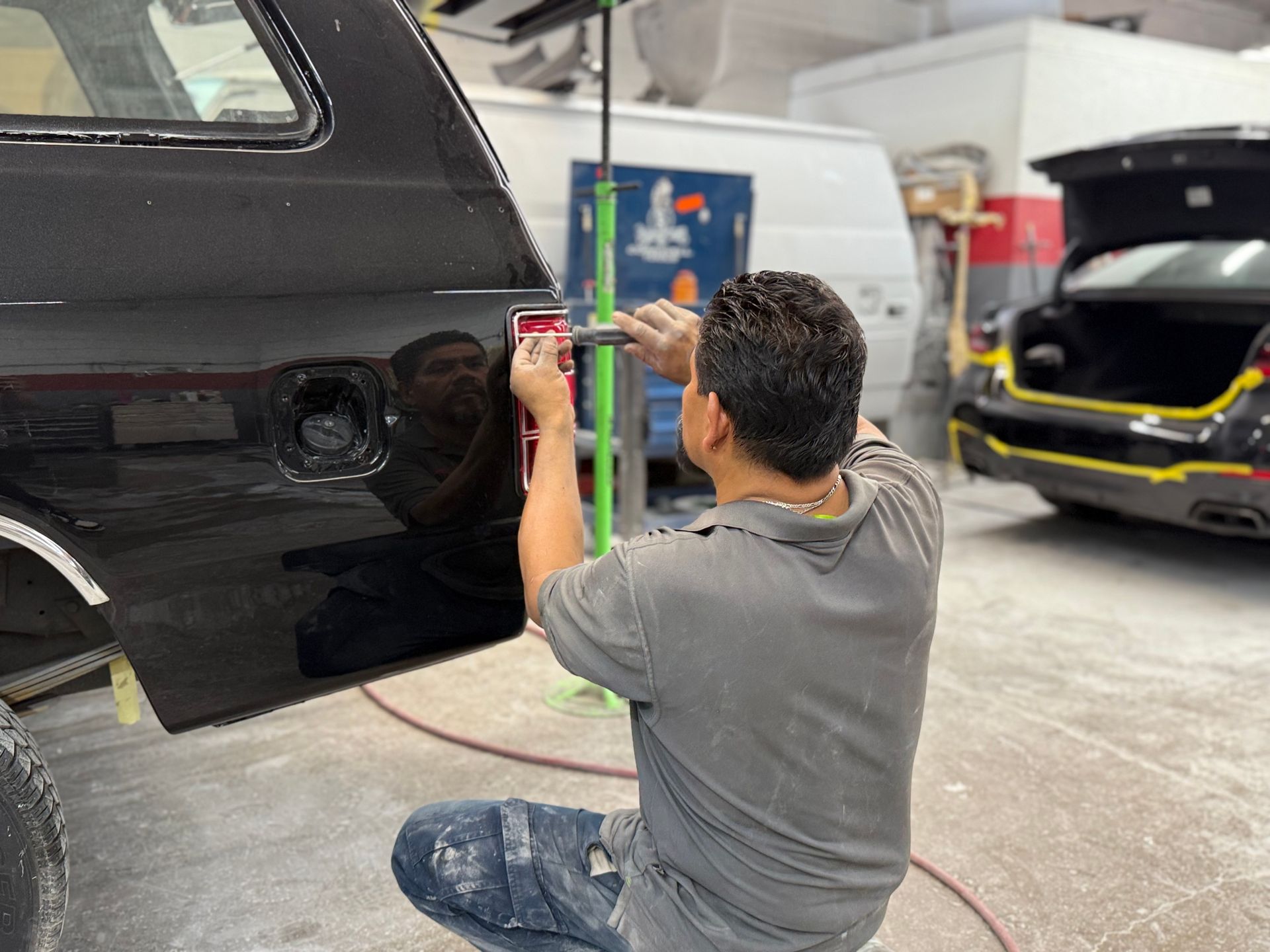 Man repairs a black car's dent in a body shop, kneeling, using a tool.