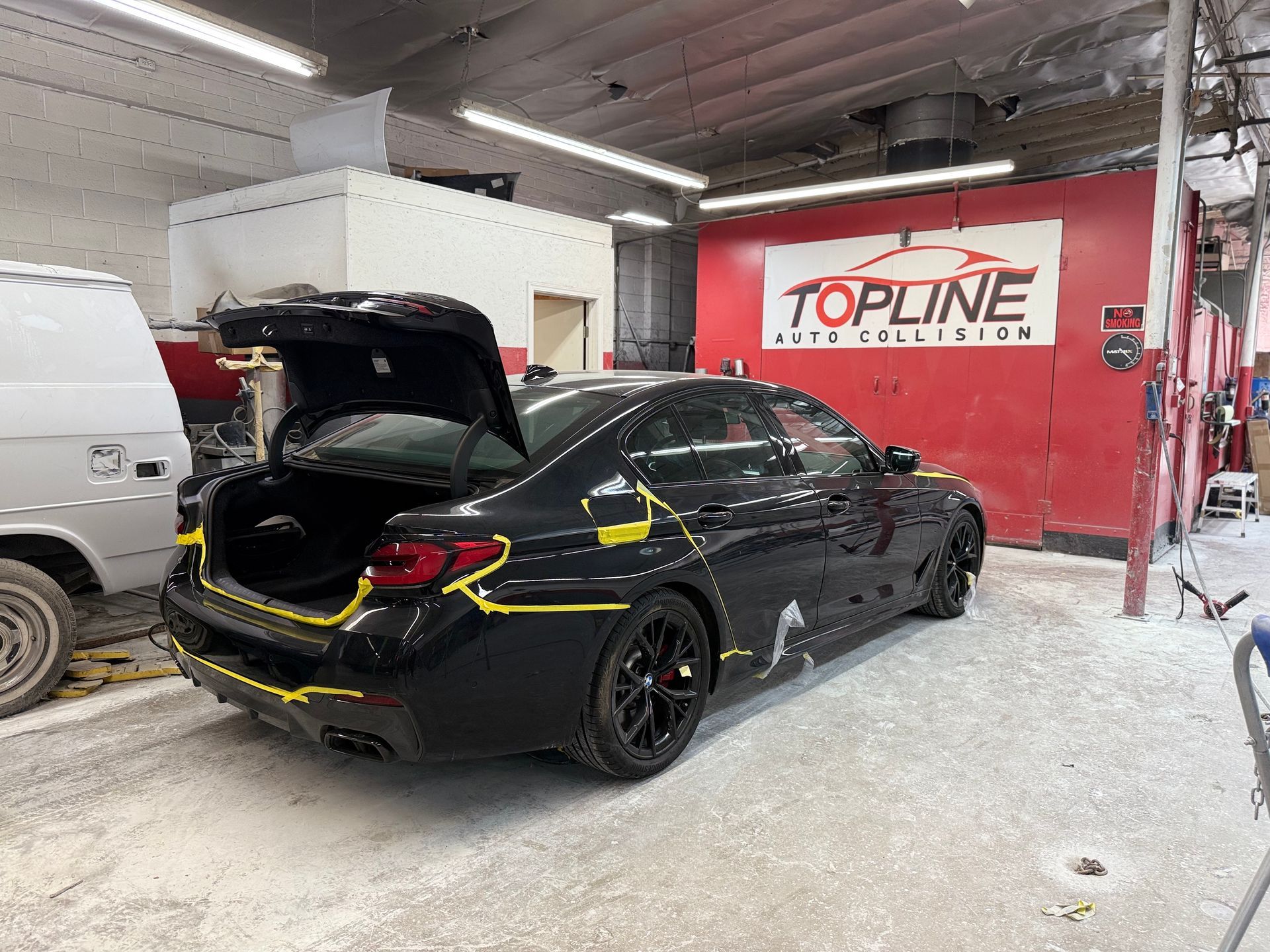 Black car in a body shop with yellow tape marking areas, Topline Auto Collision sign.