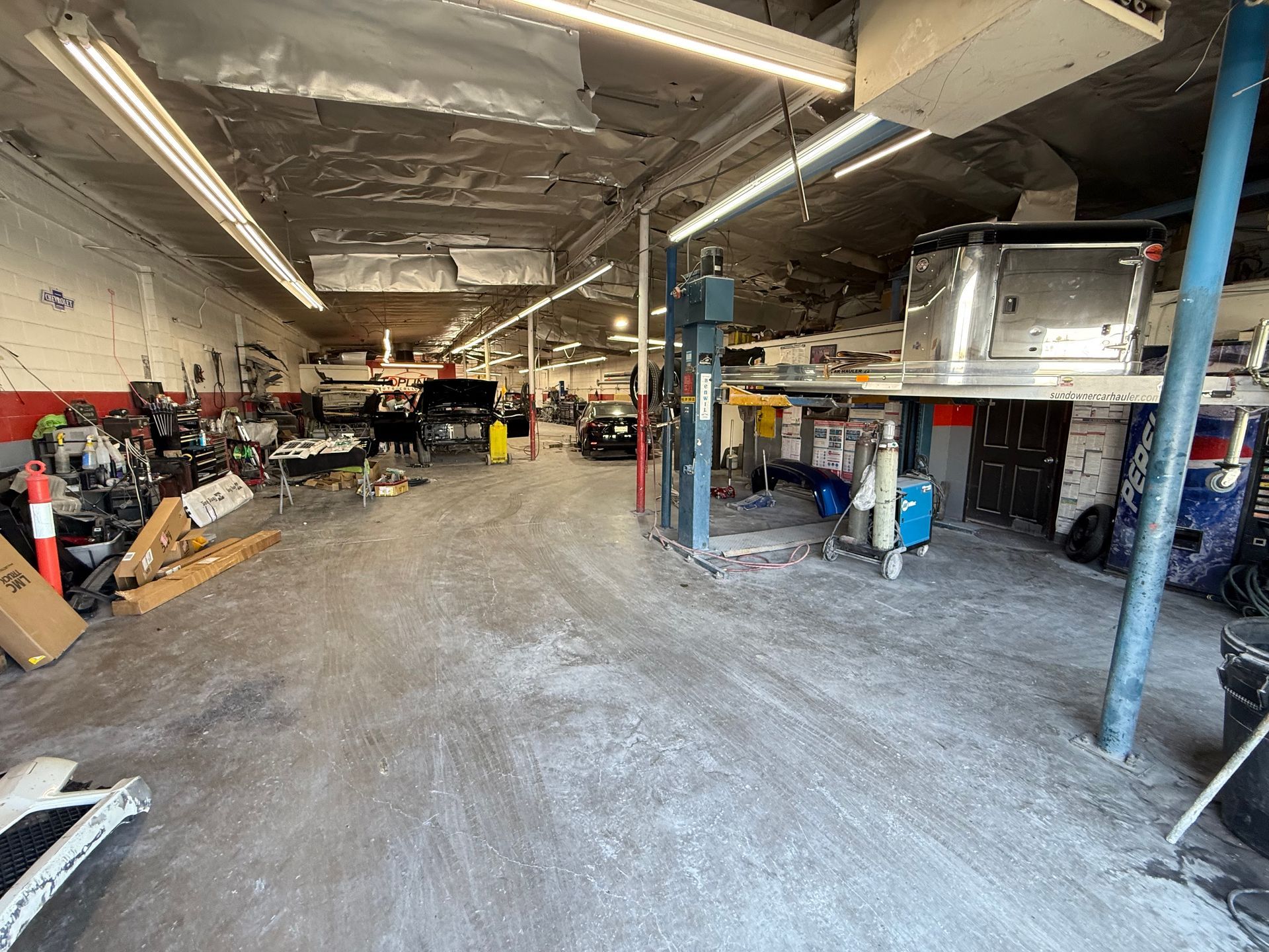 A cluttered auto repair shop interior with a car on a lift, workers, and tools.