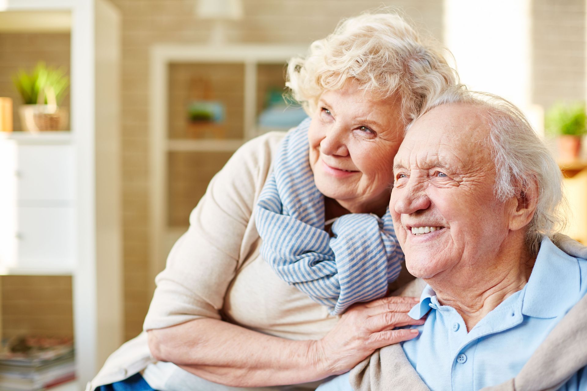 An elderly couple is hugging each other in a living room.