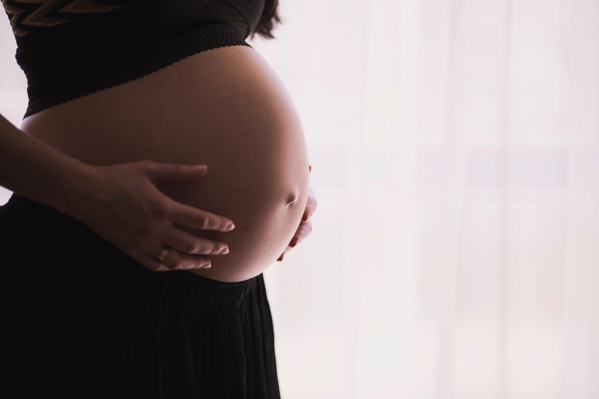 Pregnant person, side view, hands cradling large belly, dark clothing, bright background.