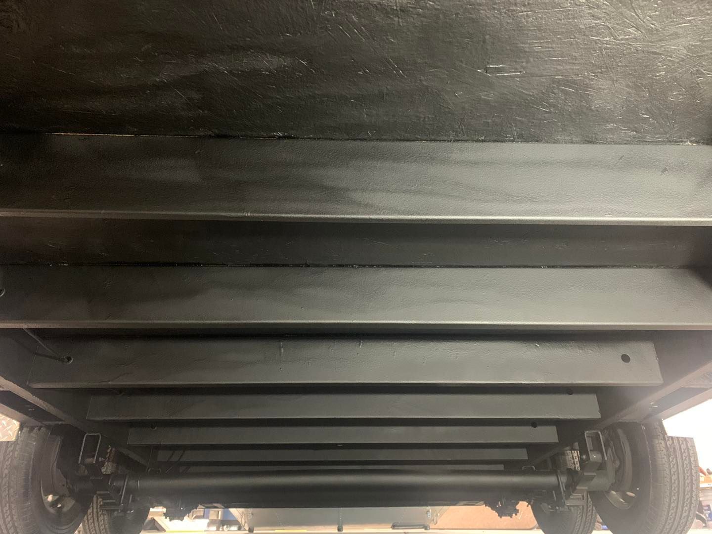 Underside view of a trailer's black steel frame. Tires are visible at the bottom.