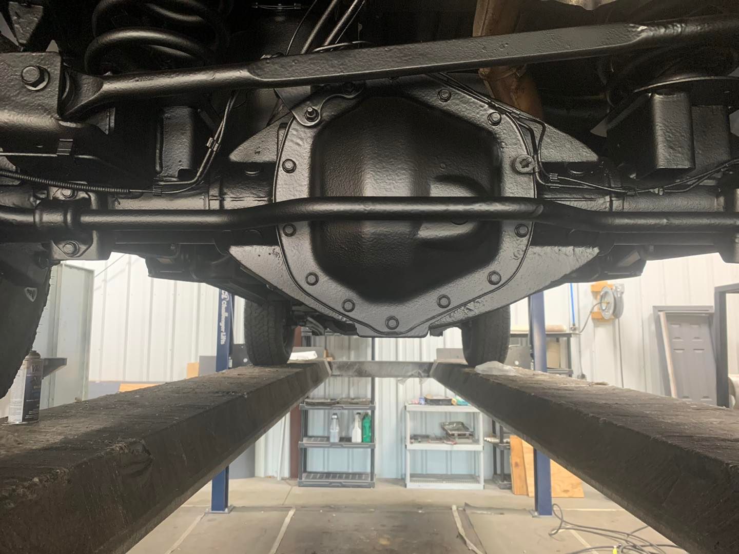 Underside of a vehicle on a lift, showing the rear axle, painted black, in a garage.
