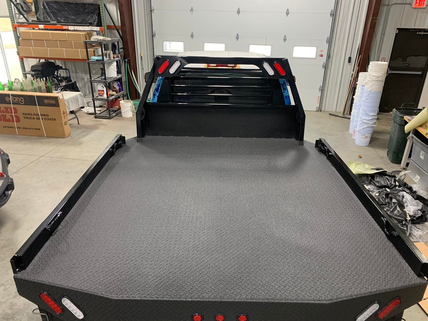 Black flatbed truck with diamond plate bed, red and white lights, and a cab protector in a garage.