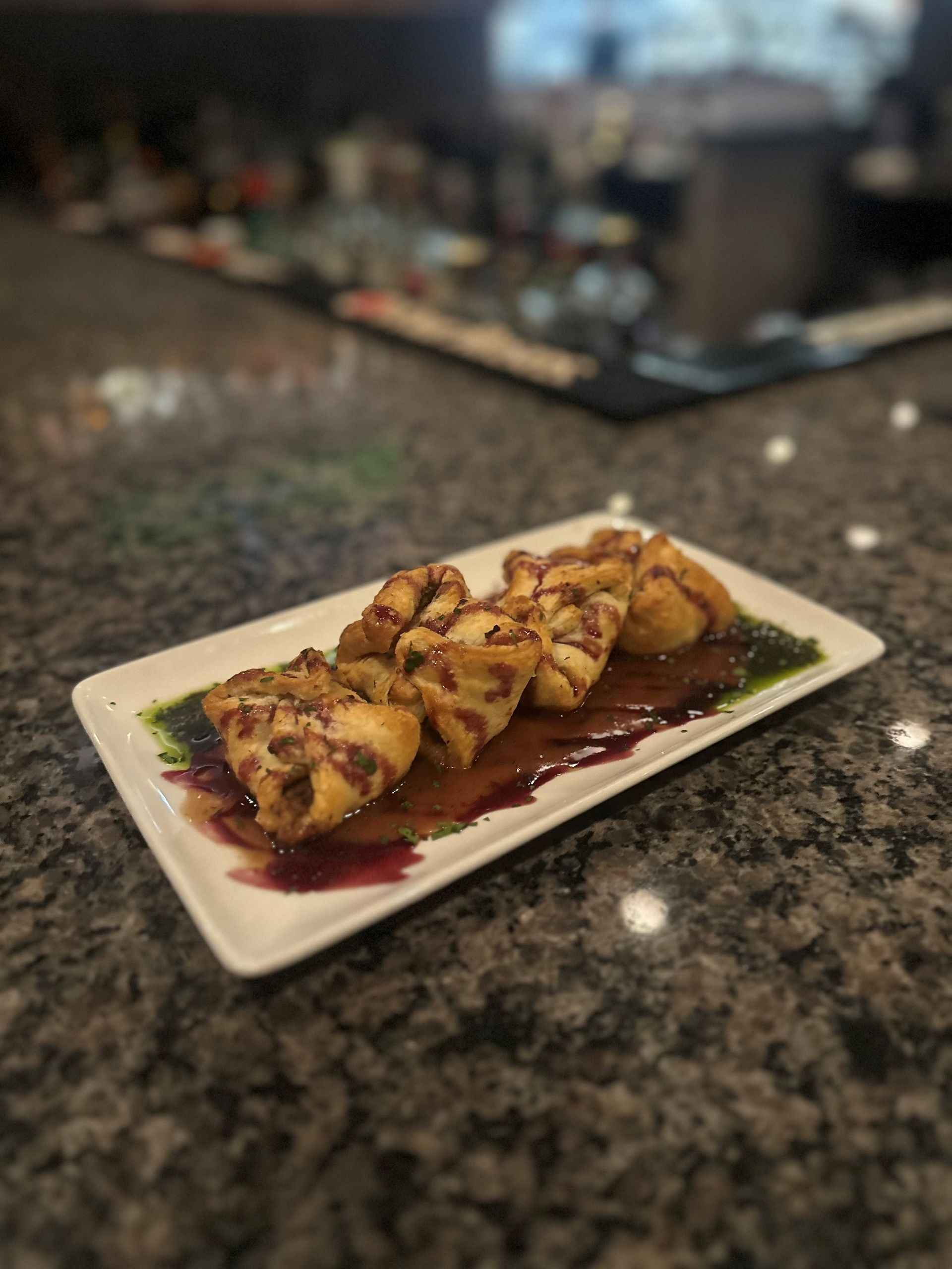 Fried wontons with sauce on a white plate on a speckled countertop, blurry bar in background.