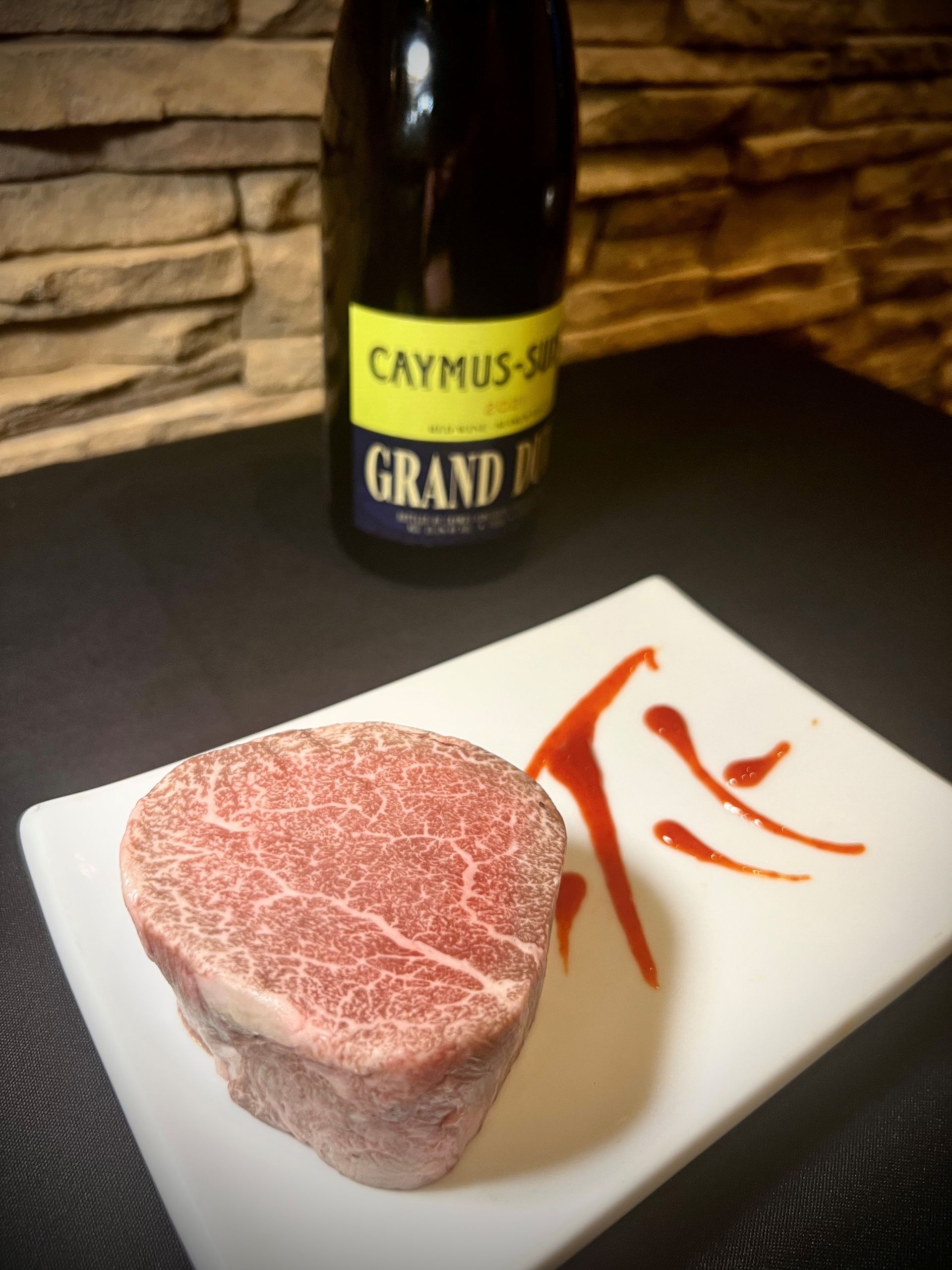 A well-marbled steak on a white plate with red sauce drizzle, Caymus-Suisun wine in the background.