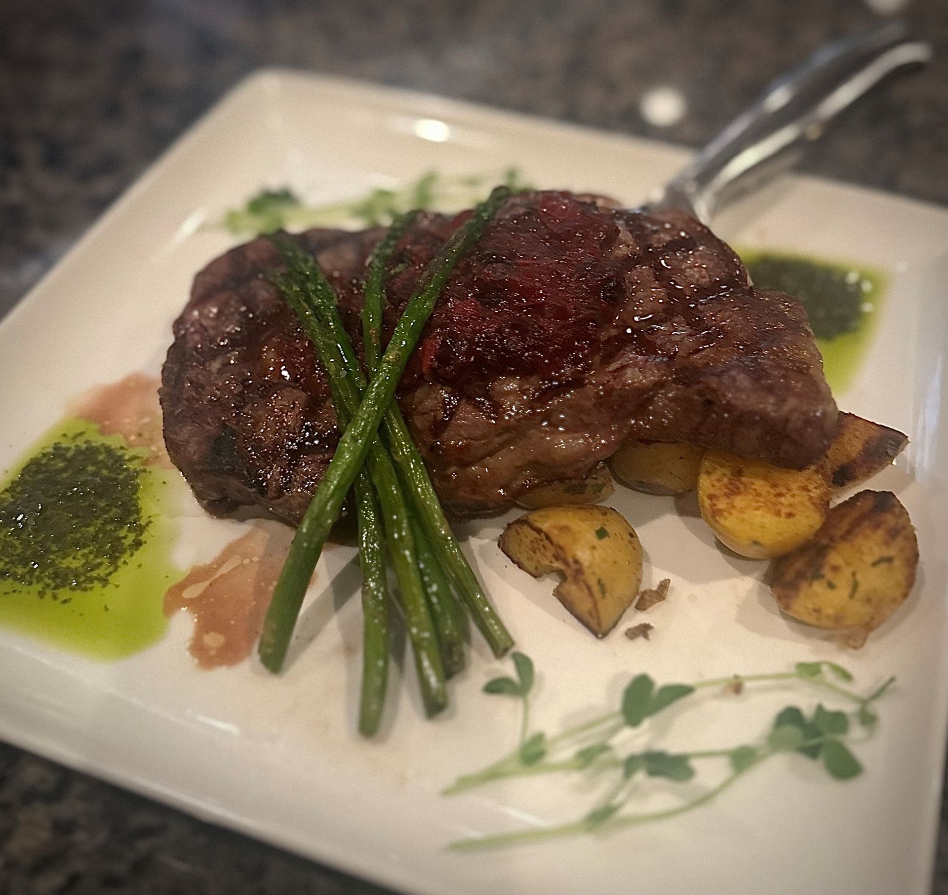 Seared steak with asparagus and potatoes on a white plate, drizzled with sauces and garnish.
