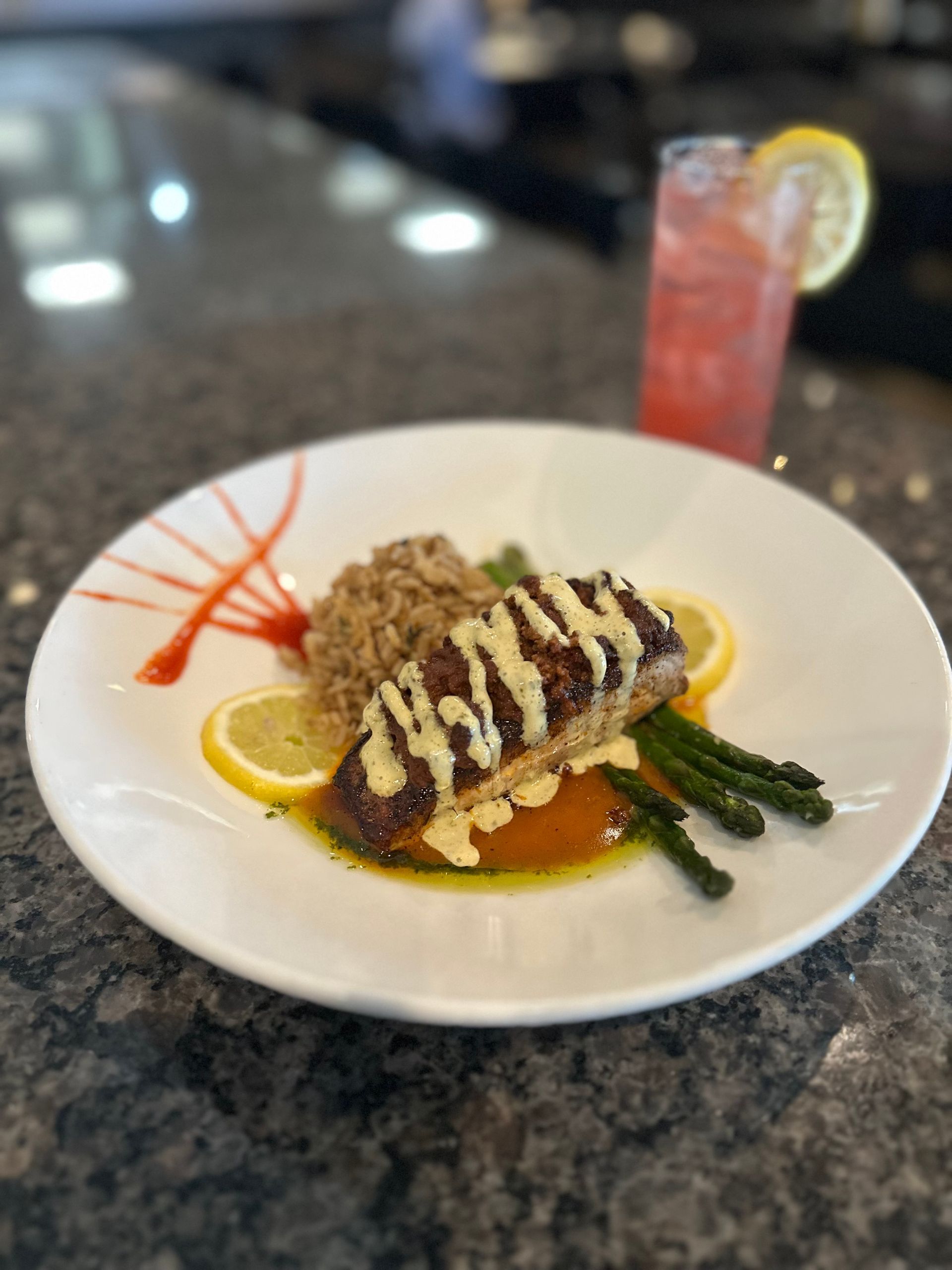 Plated entrée with grilled meat, vegetables, and sauce on a restaurant table beside a pink drink.