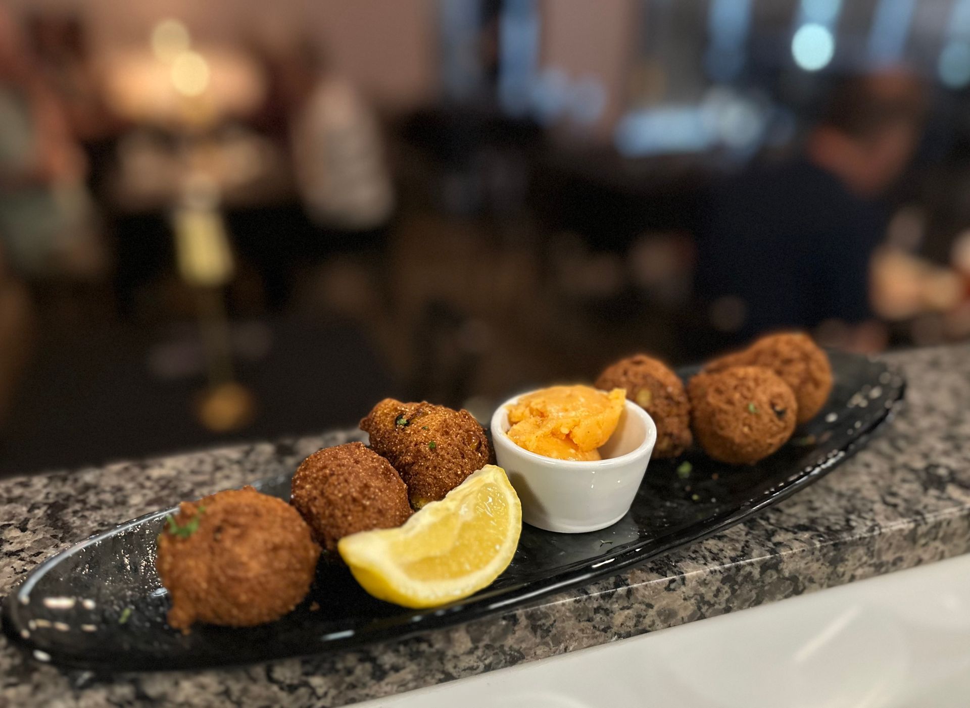 Assorted fried appetizers on a black platter with lemon wedges and dipping sauce in a restaurant.