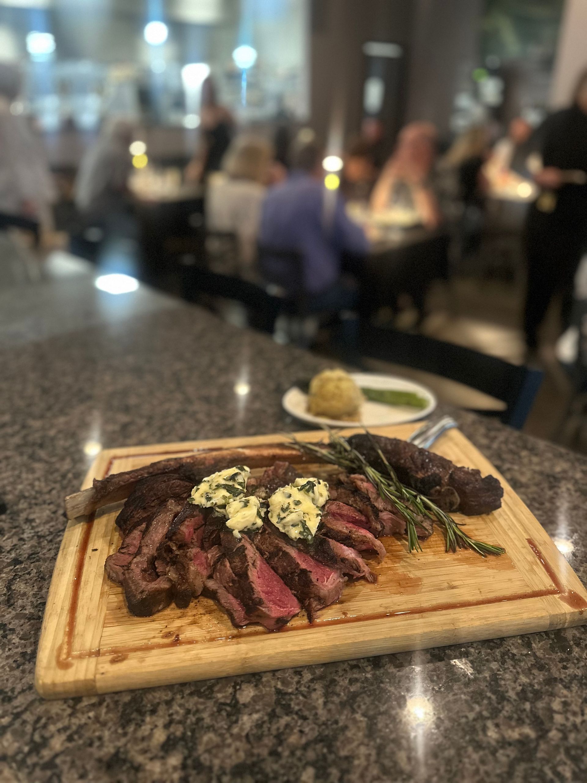Steak platter with herb butter on a wooden board at a restaurant table, with a blurred dining room background