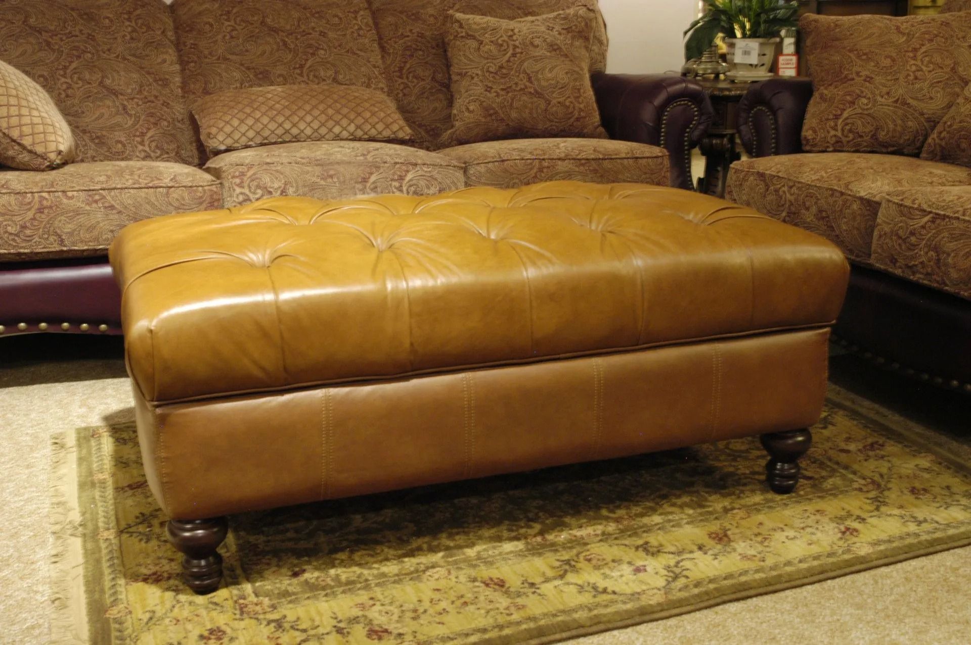 Tan leather tufted ottoman on a patterned rug in front of a matching sofa.