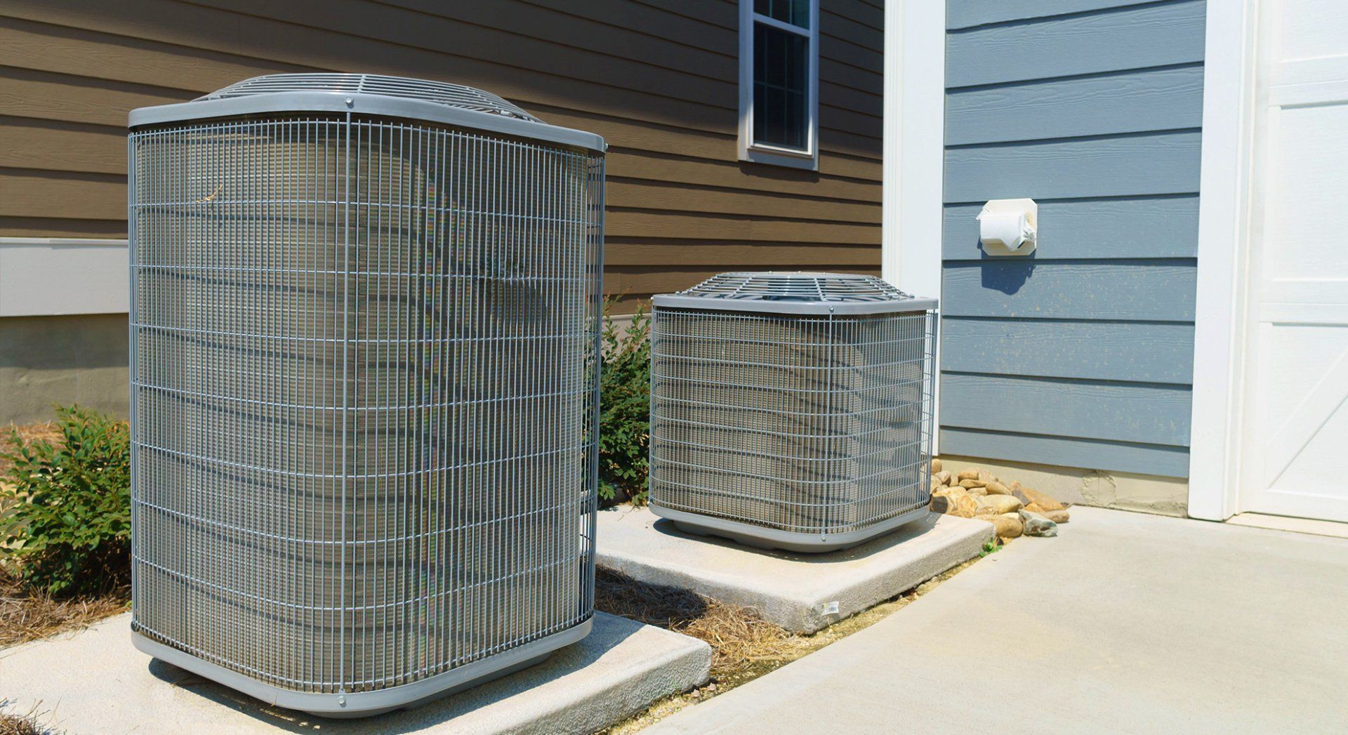 Two outdoor HVAC condenser units beside a house on a concrete patio.
