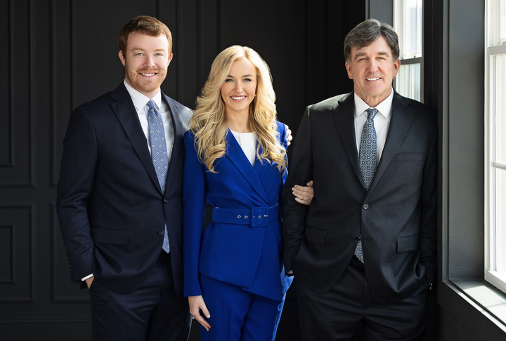 Three people in business suits standing together by a window, smiling at the camera.