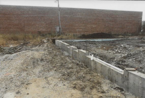 Concrete foundation with retaining wall under construction in a dirt lot, brick building in the background.