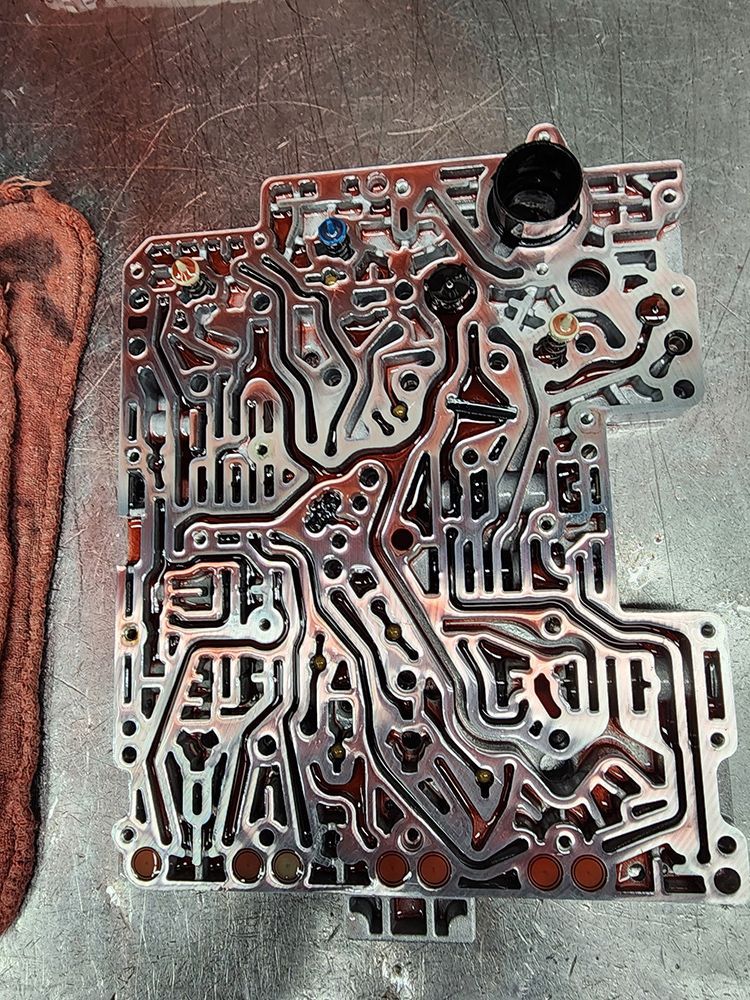 A transmission valve body with intricate channels, viewed from above, on a metal surface.