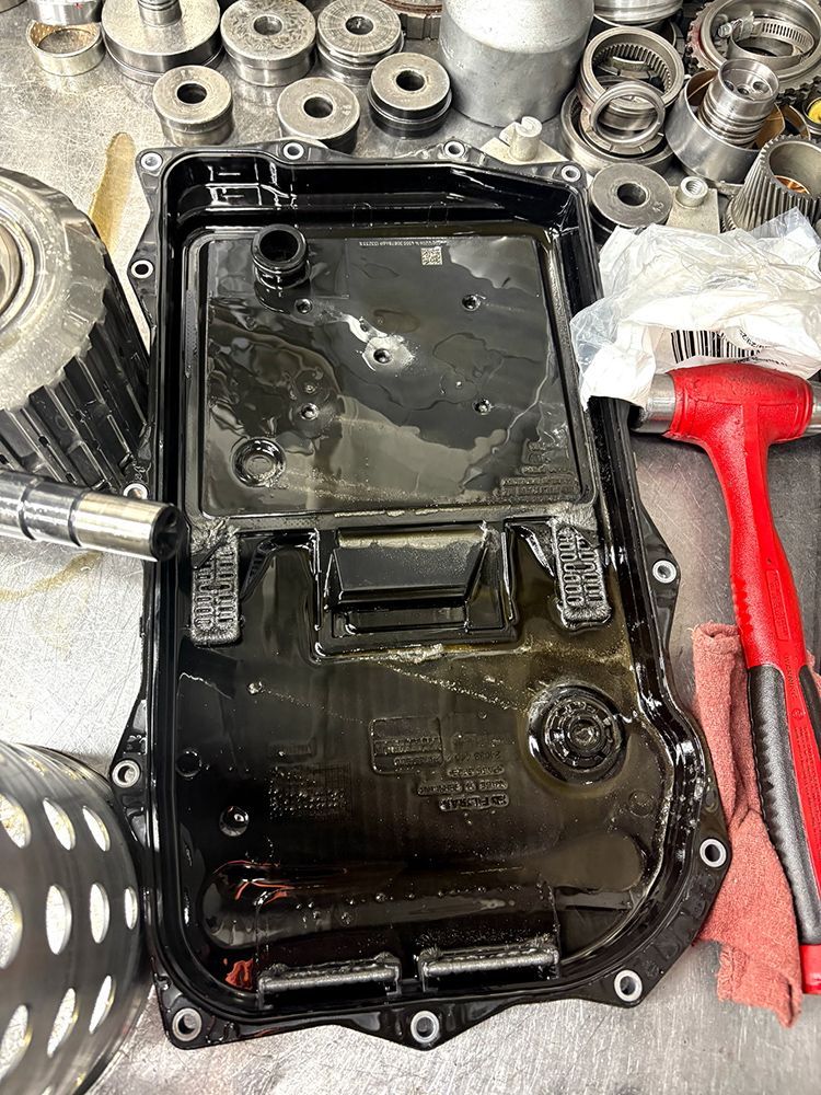 Black automatic transmission oil pan. Surrounded by transmission parts, tools, and oil.