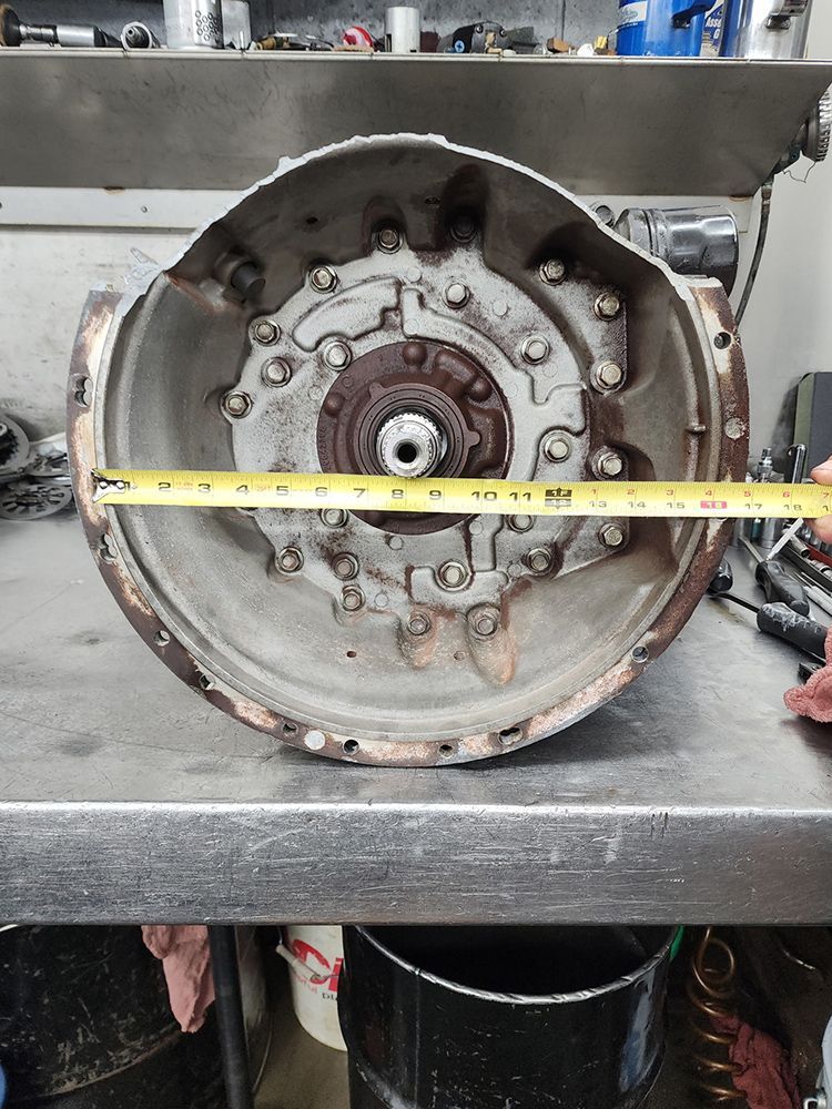 An open, circular metal component with bolts, a tape measure across it, sits on a workbench.