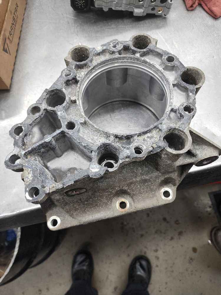 Gray metal engine component on a work surface, viewed from above, with numerous holes around a central cylinder.