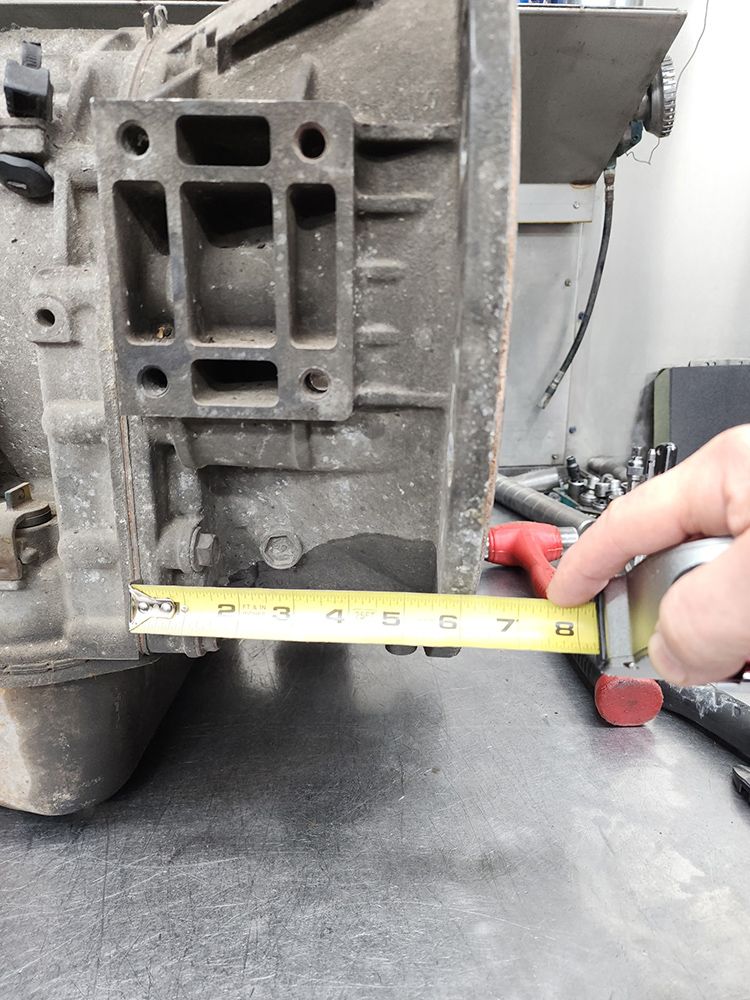 A person measures a metal transmission part with a yellow tape measure in a workshop.