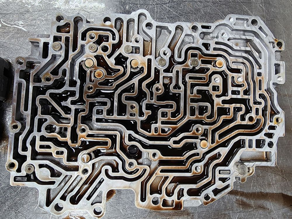 Metallic transmission valve body with intricate channels, coated in dark fluid.