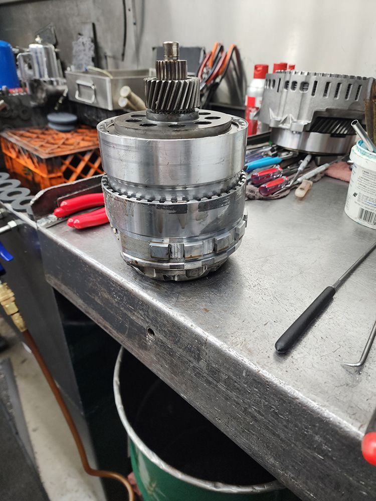 Transmission component assembly on a workbench. Metallic parts are stacked, gear at the top. Tools and workshop setting.