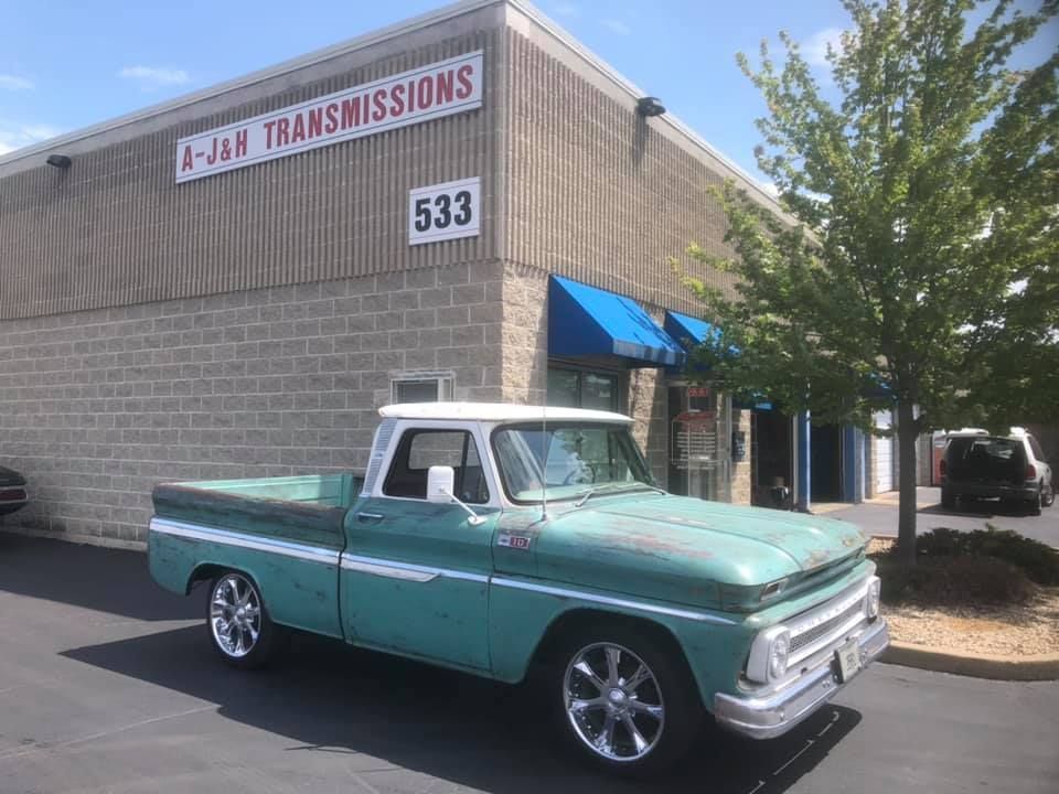 Teal classic pickup truck parked outside A-J-H Transmissions shop, brick building with blue awning.