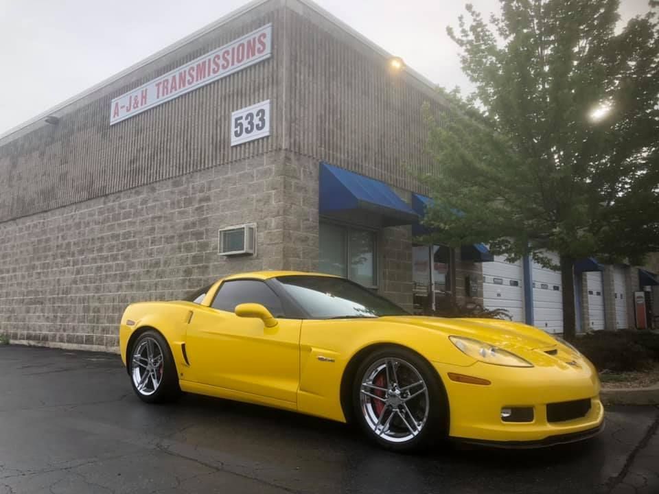 Yellow Corvette parked in front of a transmission shop with a wood-paneled facade.