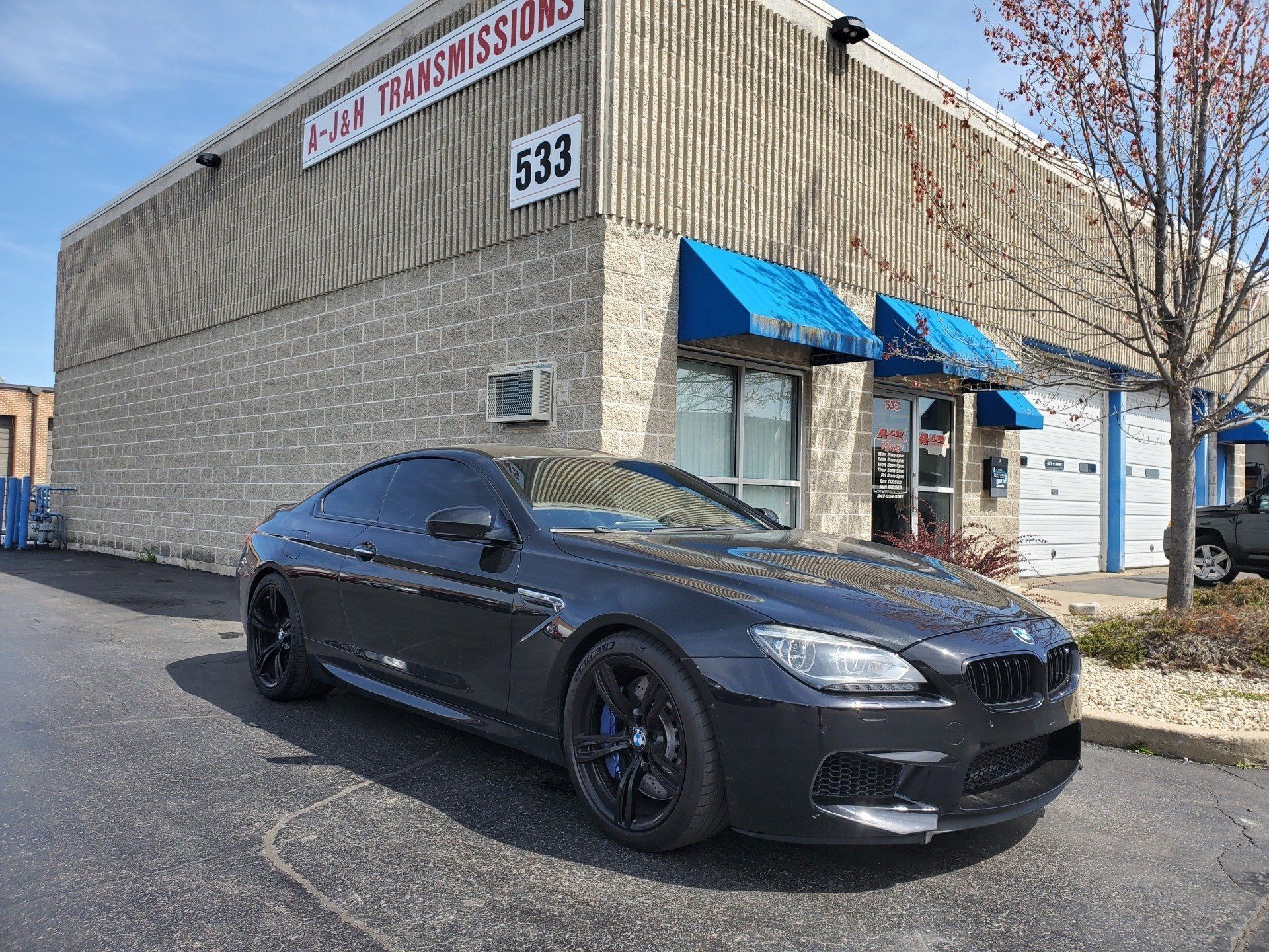 Dark gray BMW coupe parked in front of a building with a blue awning. The building is brick and says 