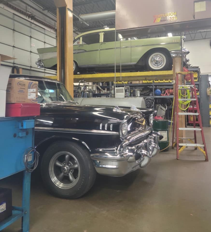 Two classic cars in a garage: a black one in front, a green one raised on a lift. Tools and equipment surround.