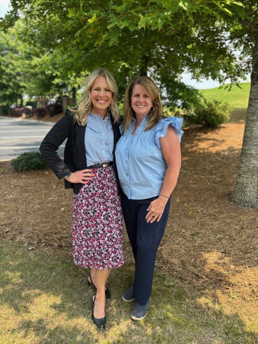 Two women standing outside, smiling. One in a floral skirt and black jacket, the other in a blue top and pants.