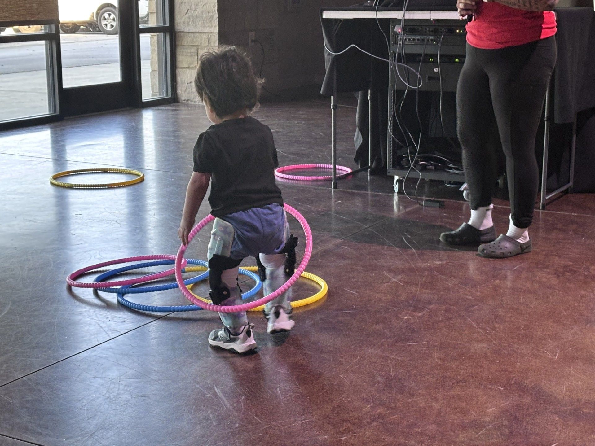 Child walking among colorful hoops on a gym floor, with an adult standing nearby.