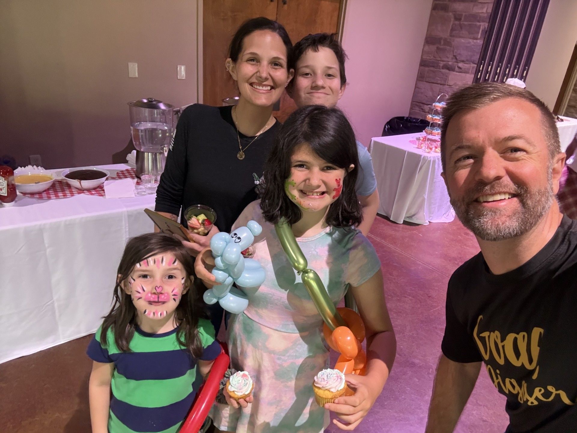 Five people smiling for a selfie at a party, with children holding colorful balloons and stuffed animals.