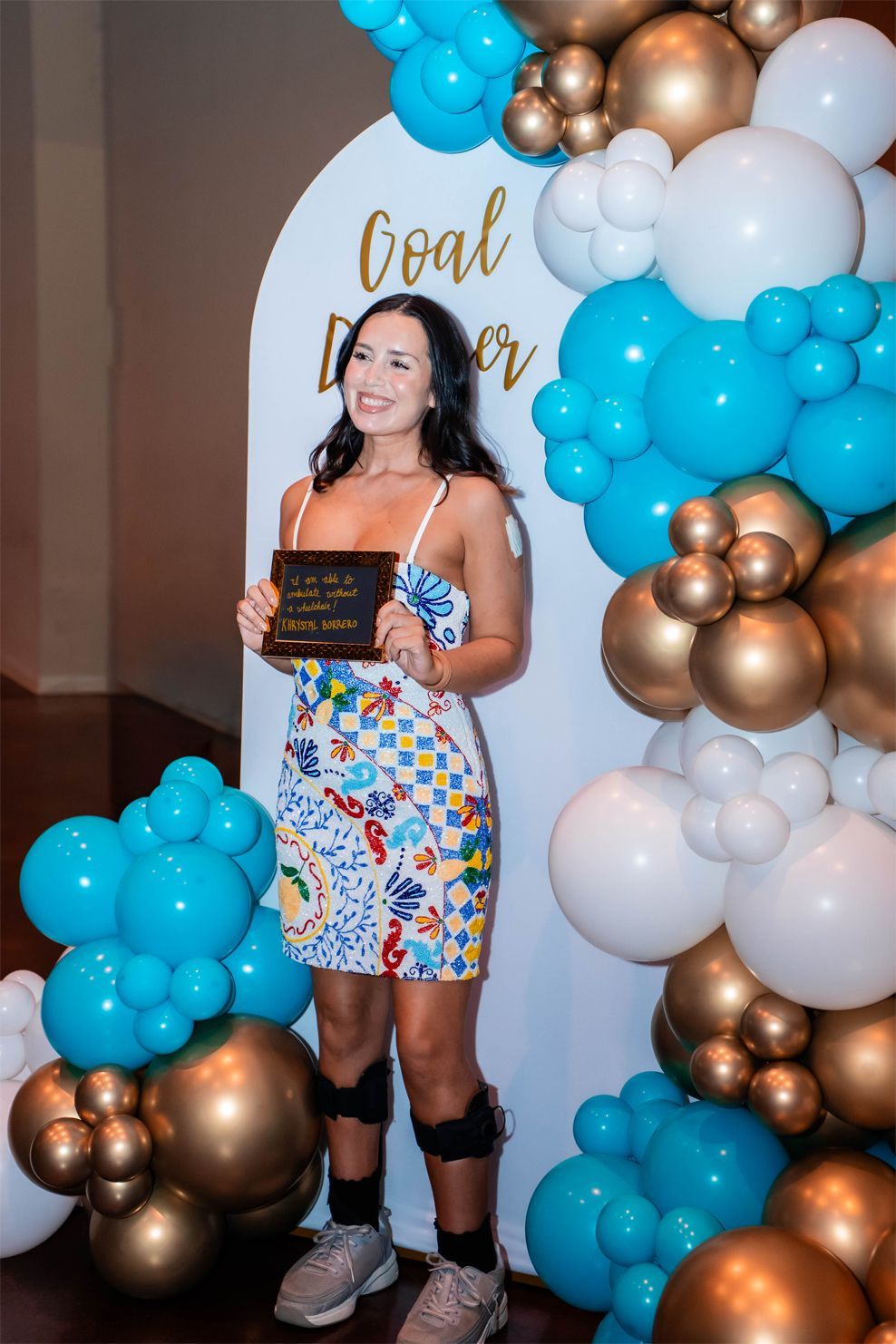 A person smiles holding an award in front of a blue, gold, and white balloon arch with Goal Digger on the backdrop.
