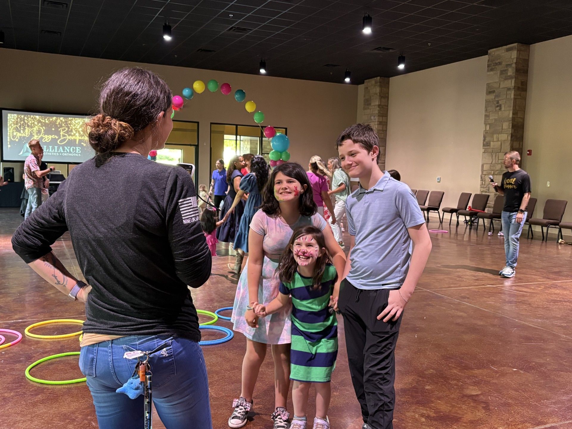 Woman posing for a photo with two smiling children in a large indoor event hall with colorful hoops on the floor