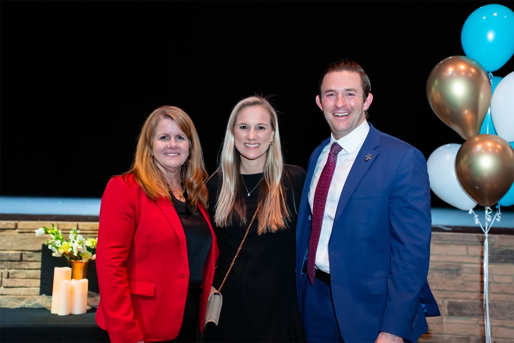 Three people smiling together in formal attire at an event with gold and blue balloons in the background.