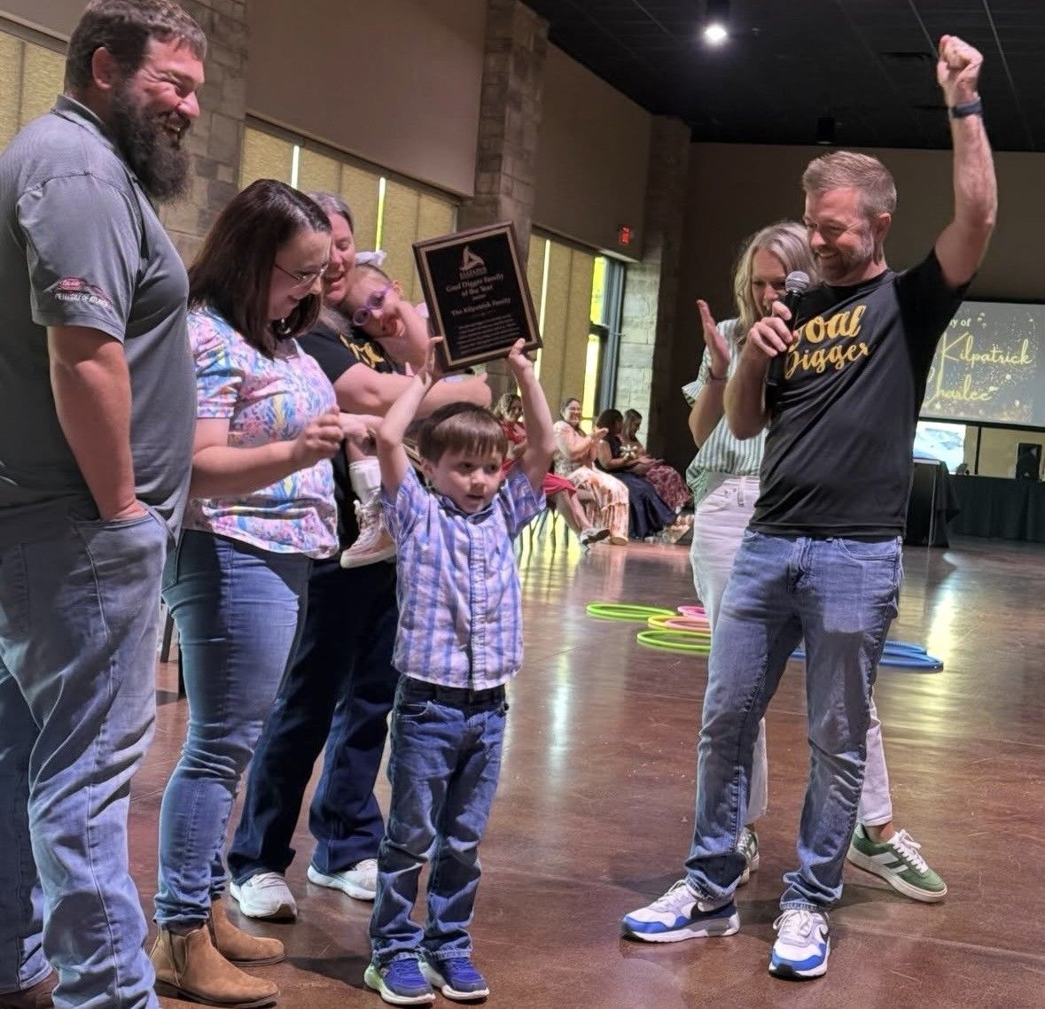 People celebrating indoors, with a child holding a plaque while two adults cheer nearby